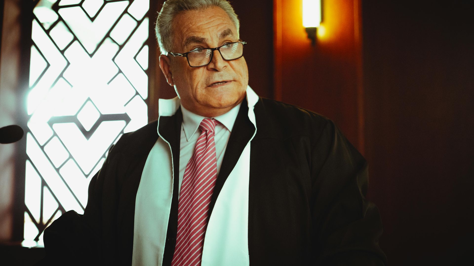 A male judge in a courtroom in Baghdad wearing a robe and tie, in a contemplative pose.