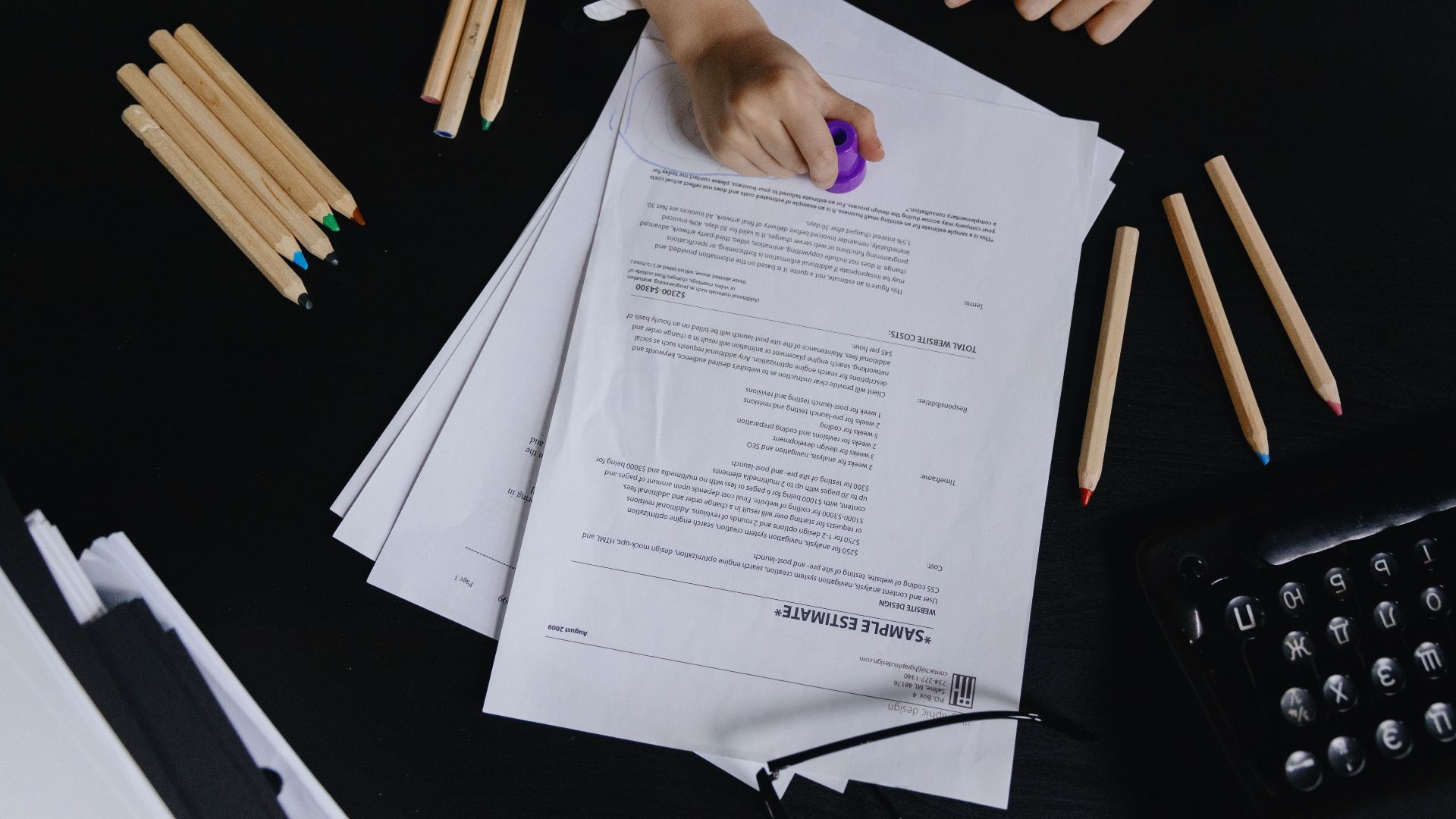 Hands working with documents and colored pencils on a black desk view.