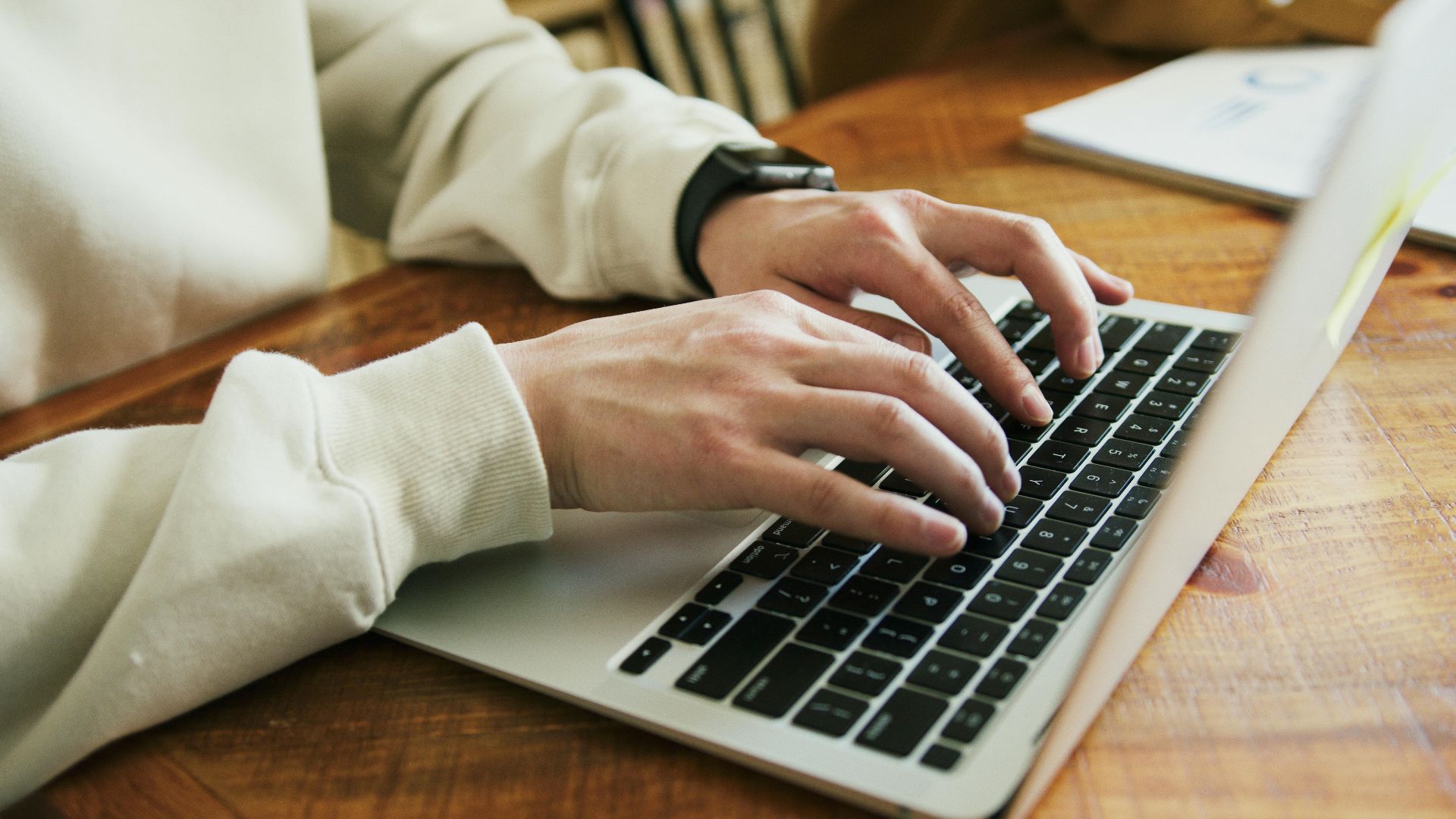 Hands typing on a laptop keyboard on a wooden desk, close-up view.