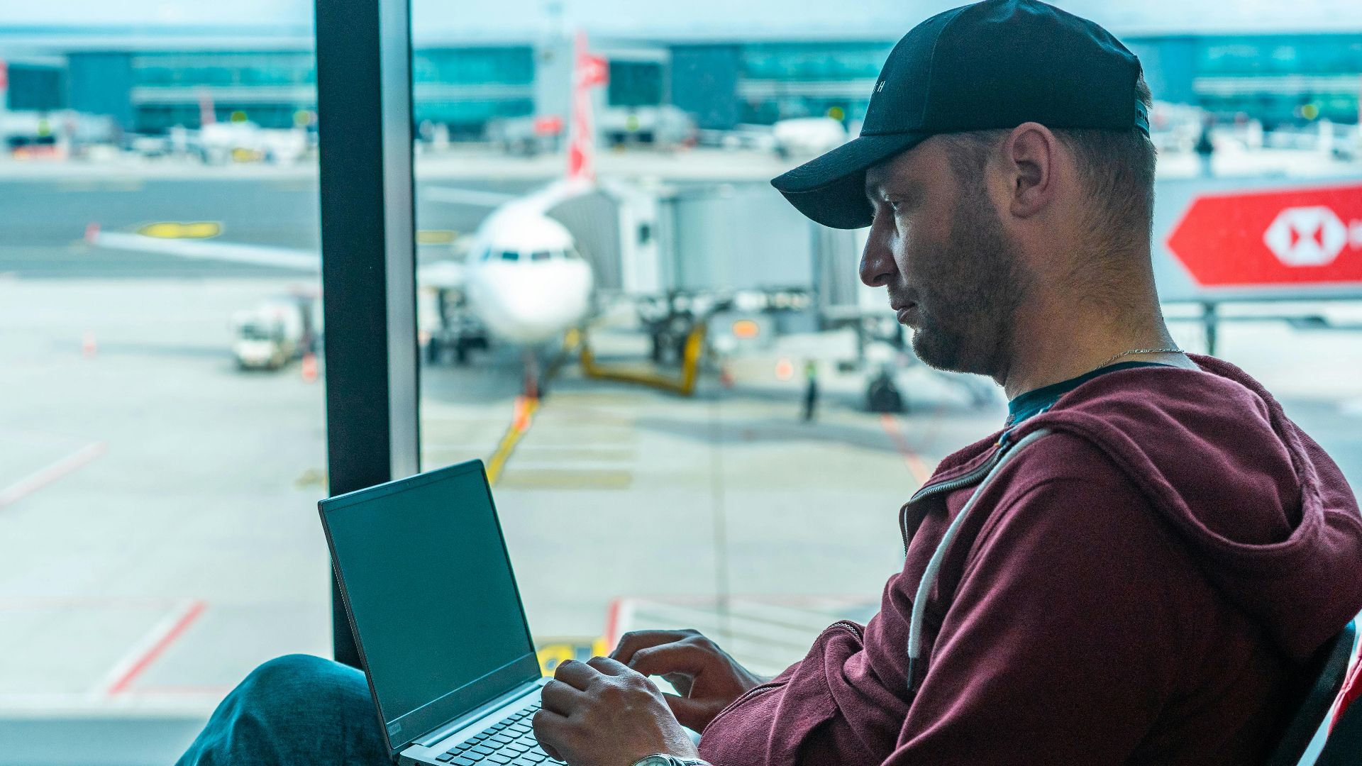 A man working on his laptop in an airport terminal, with a plane visible outside.