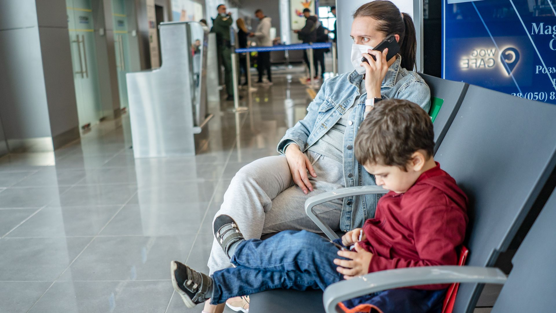 A mother and child sitting at an airport terminal. The mother is on the phone while the child is using a device.