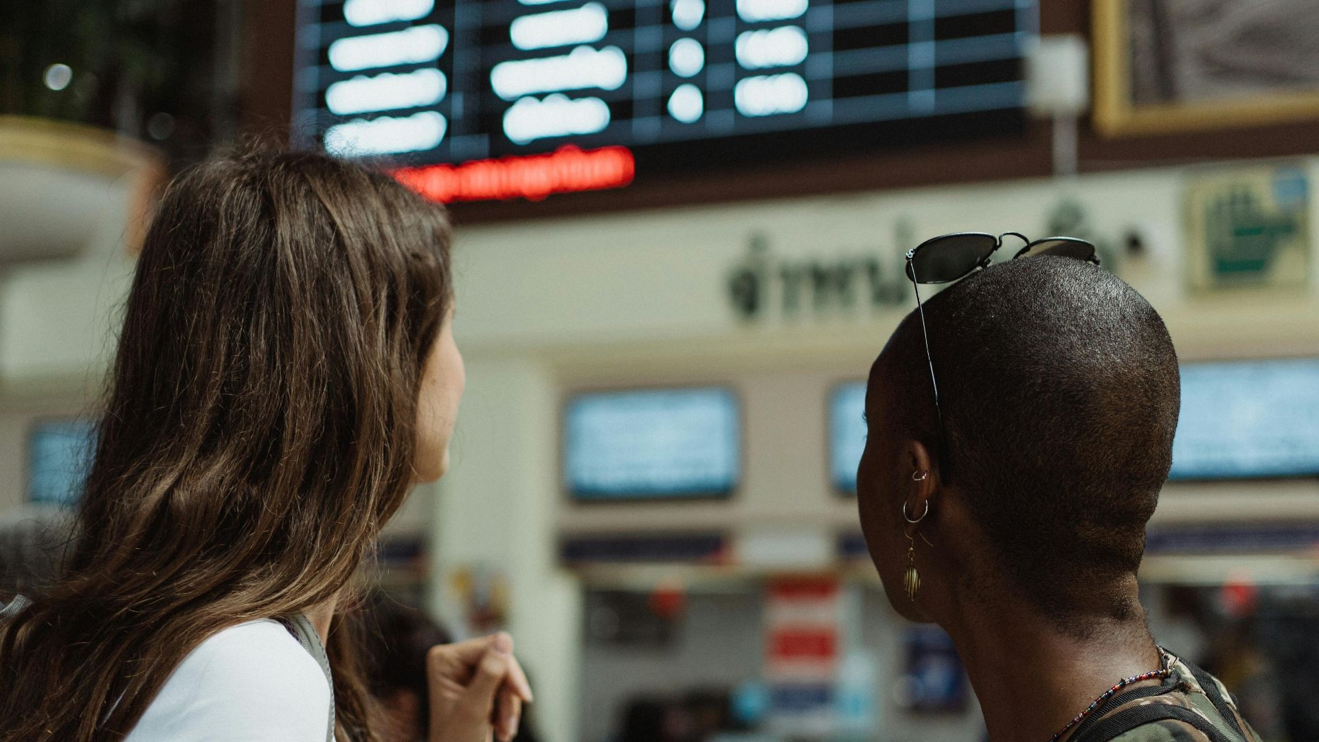 Two women checking the flight schedule on a departure board in an airport terminal, preparing for travel.