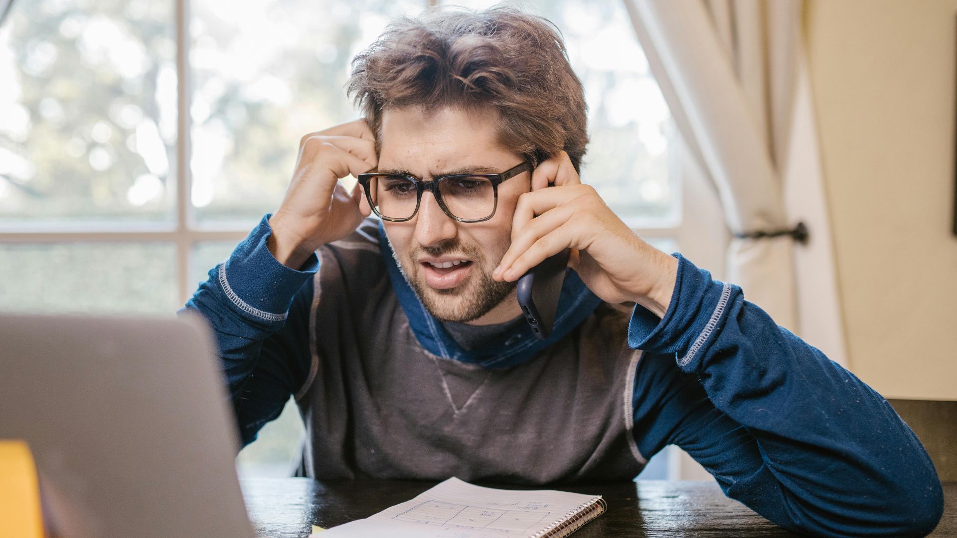 Man in glasses using laptop and smartphone at home, appearing confused.