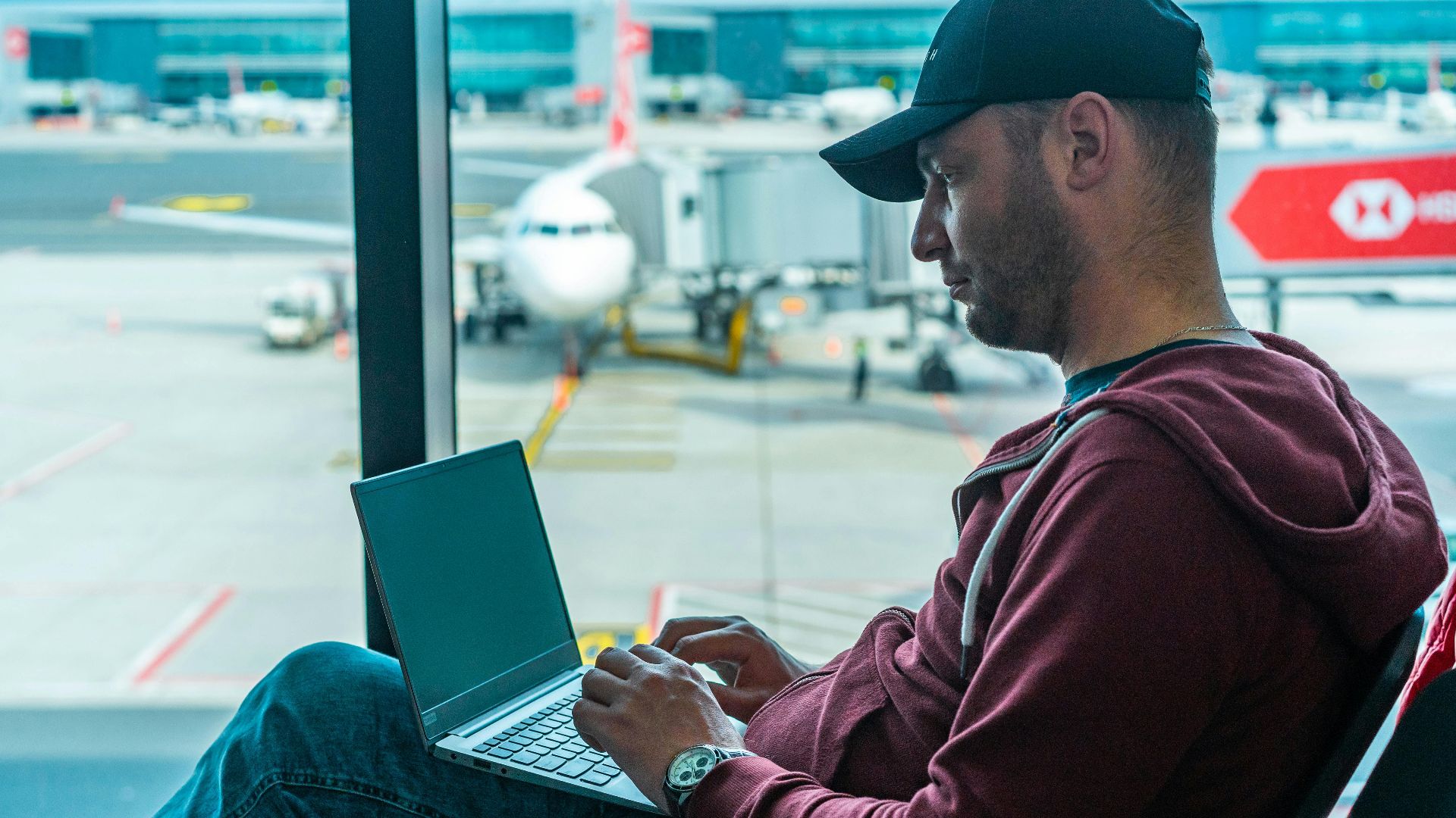 A man working on his laptop in an airport terminal, with a plane visible outside.
