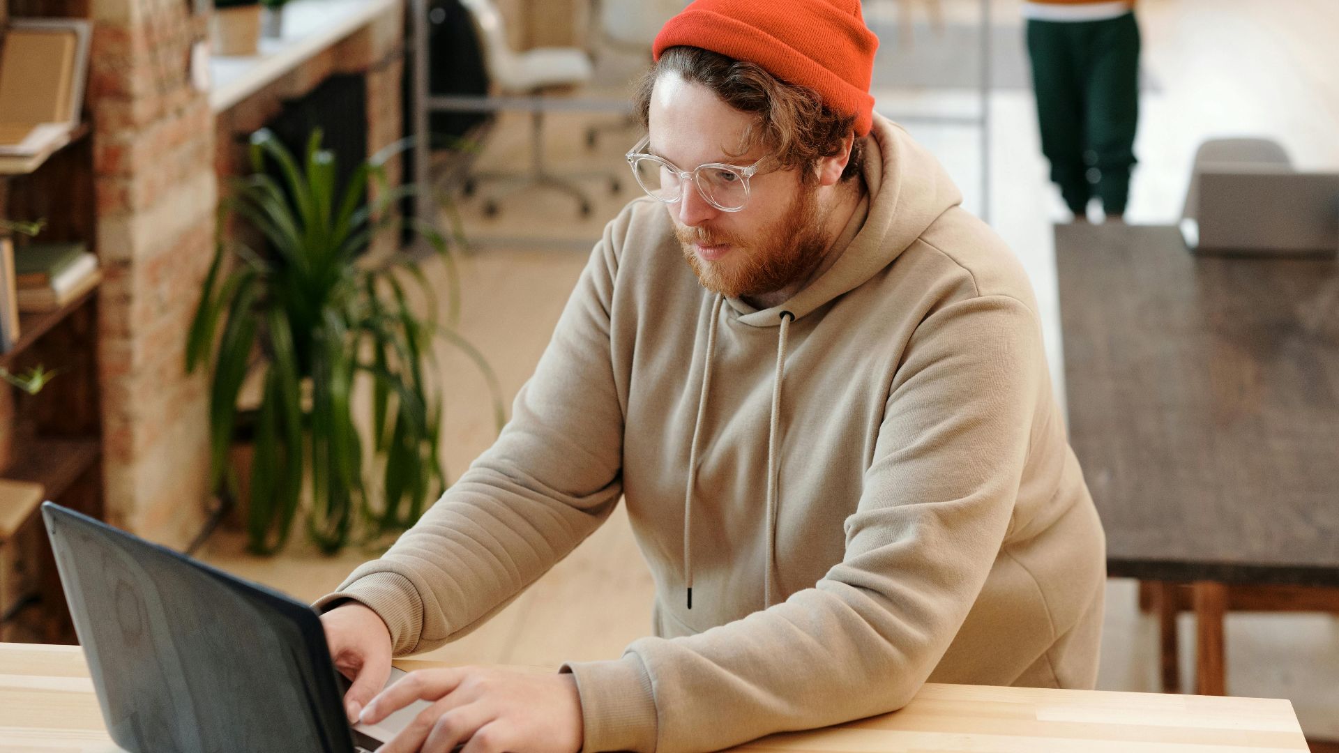 A bearded man in a hoodie works on a laptop in a modern, casual office setting.