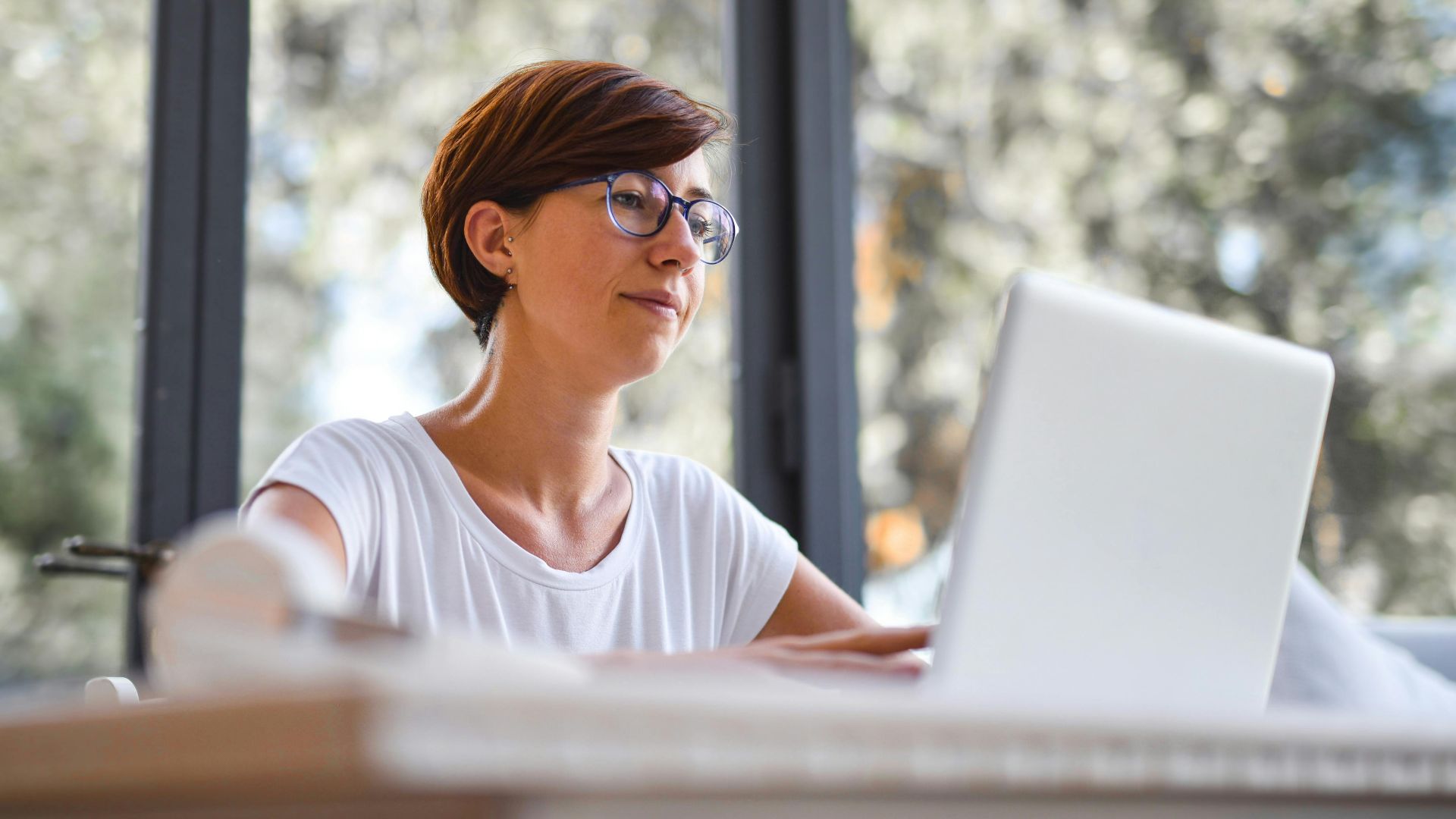 Woman with glasses using laptop in a bright home office, enjoying a peaceful work environment with natural light.