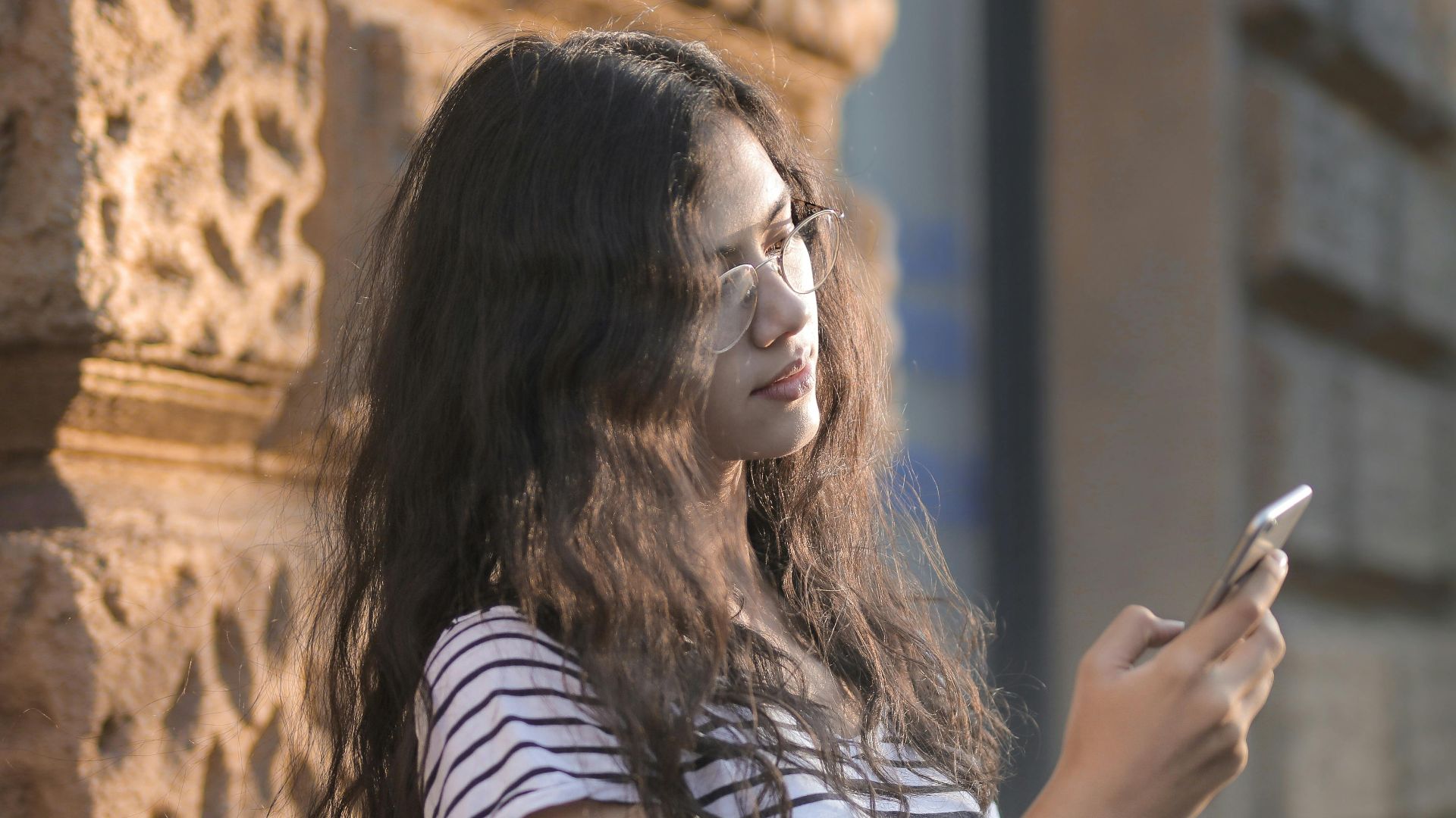 Casual portrait of a young woman leaning on a wall, using her smartphone and holding books outdoors.