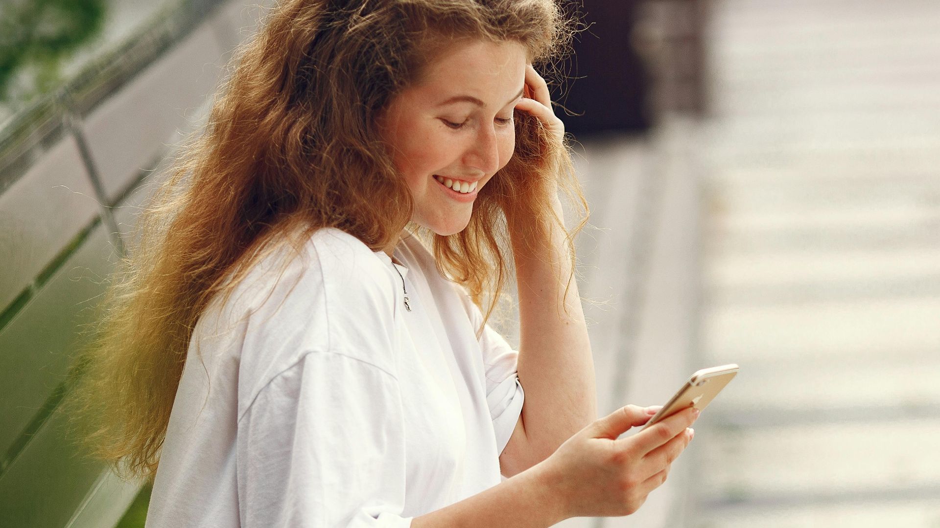 Happy young woman sitting on bench using smartphone outdoors, smiling joyfully.