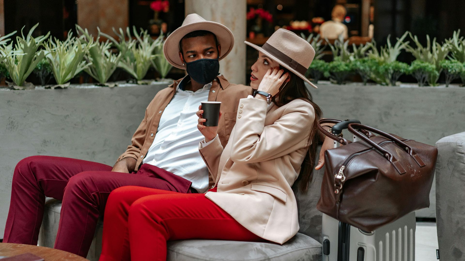 Fashionable couple seated with luggage, enjoying coffee in a chic indoor café setting.