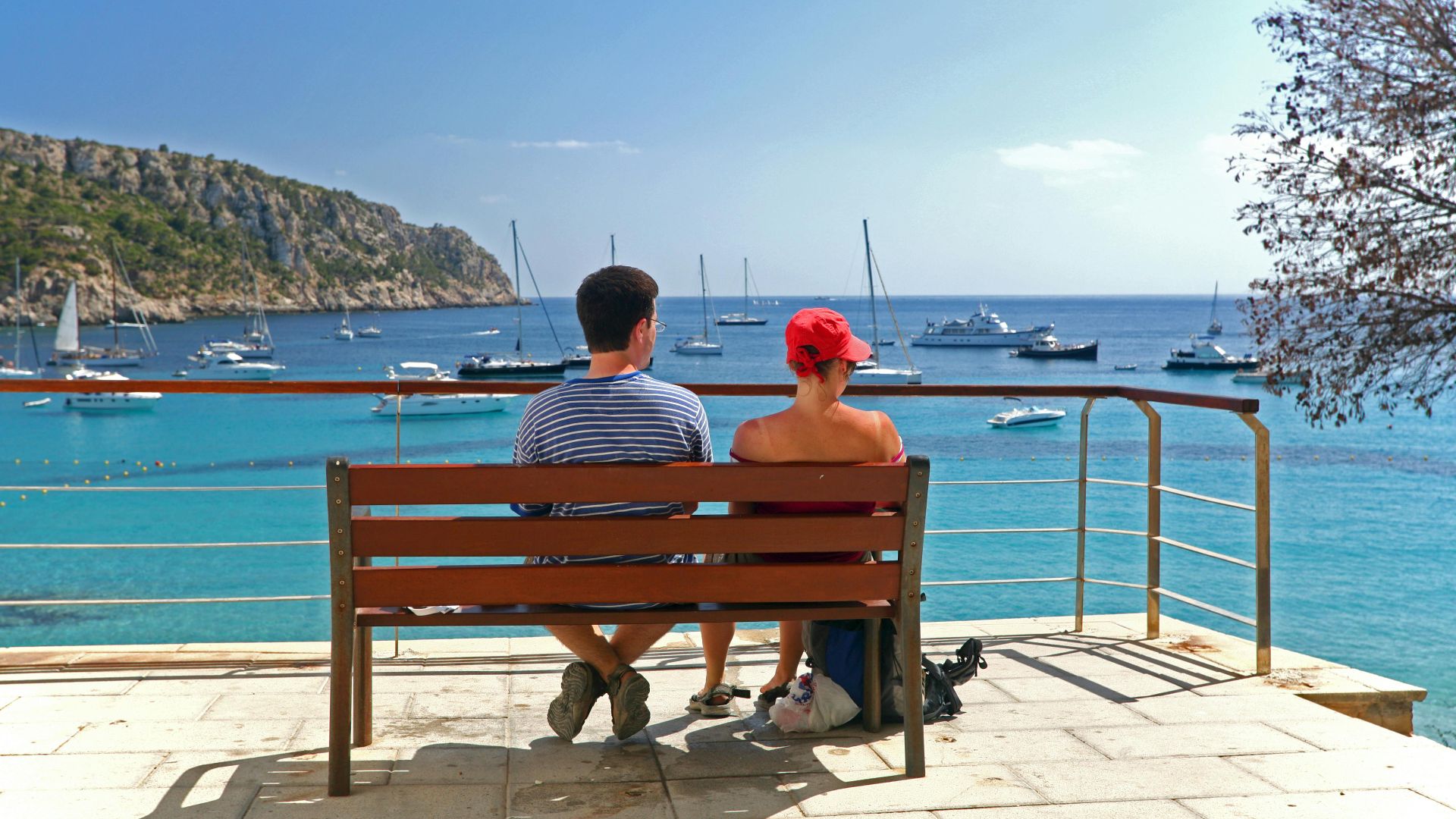 Couple sitting on a bench enjoying a scenic ocean view with boats in Ibiza, Spain.