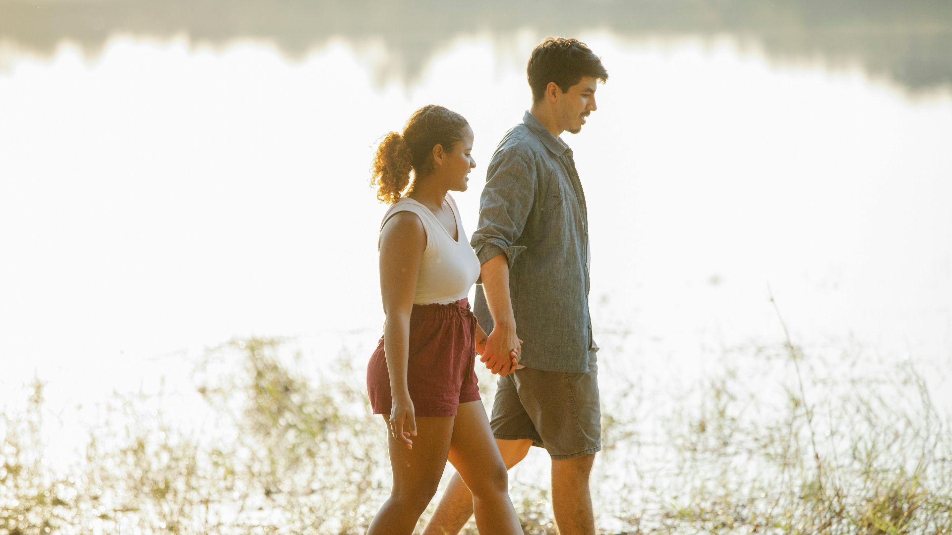 Side view of beloved young multiracial couple walking and holding hands during romantic date near lake at sunset