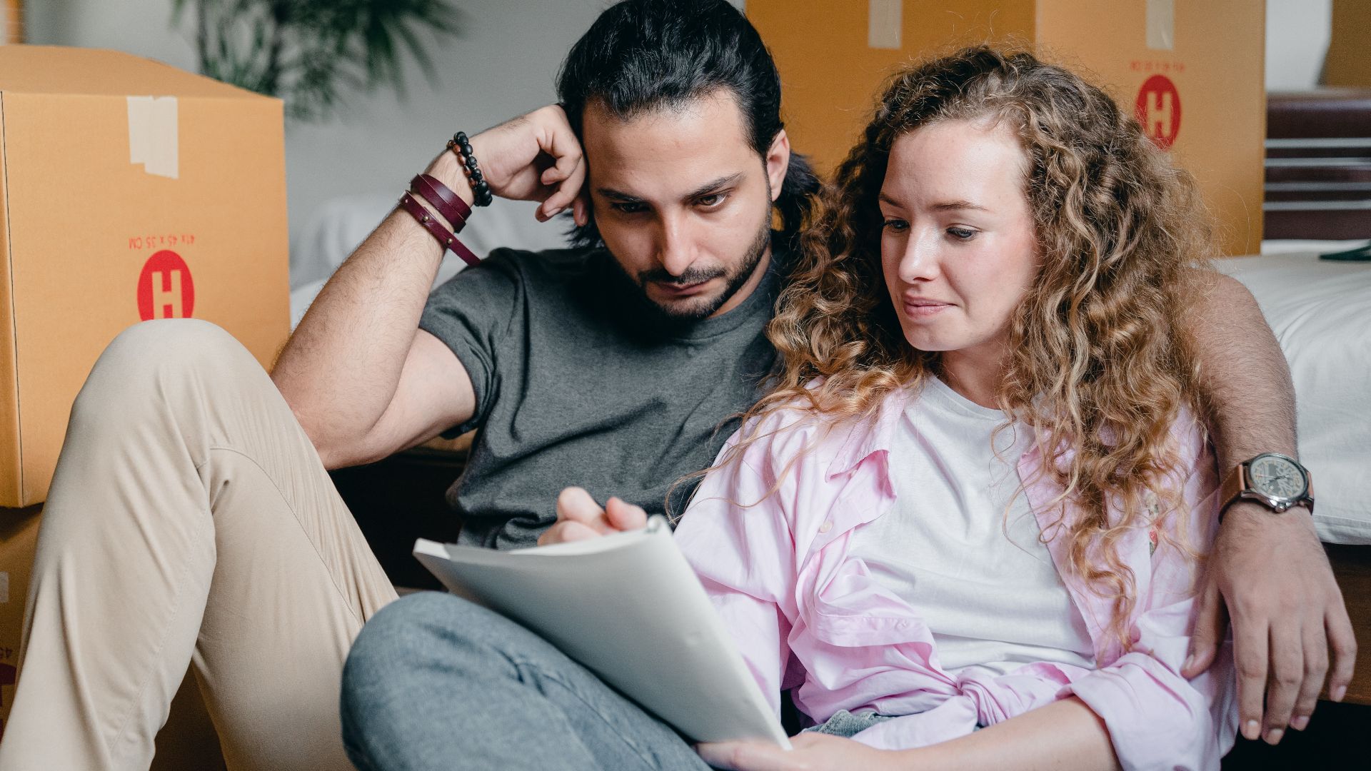 Pensive male and female in casual clothes sitting together among boxes and writing notes in notebook while leaning on bed