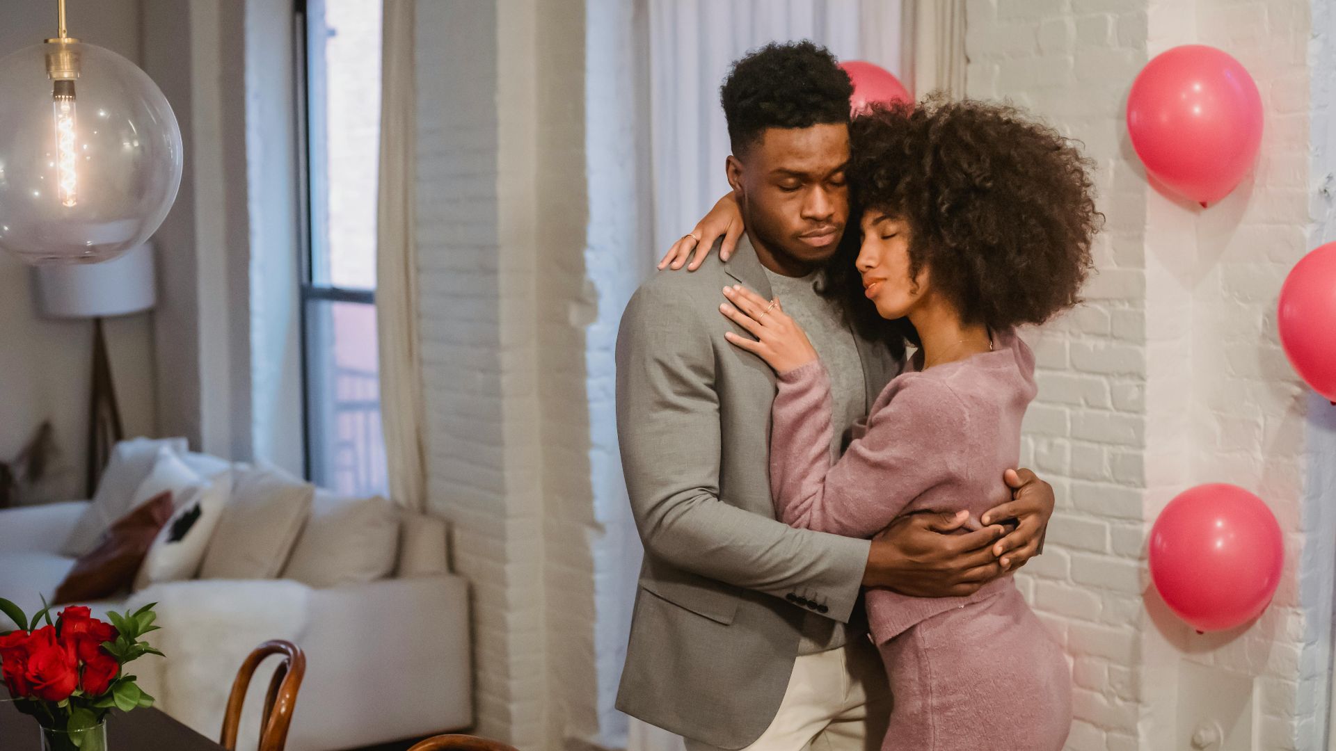 Dreamy young black couple in elegant clothes standing in light room and hugging while dancing together at brick wall with pink balloons