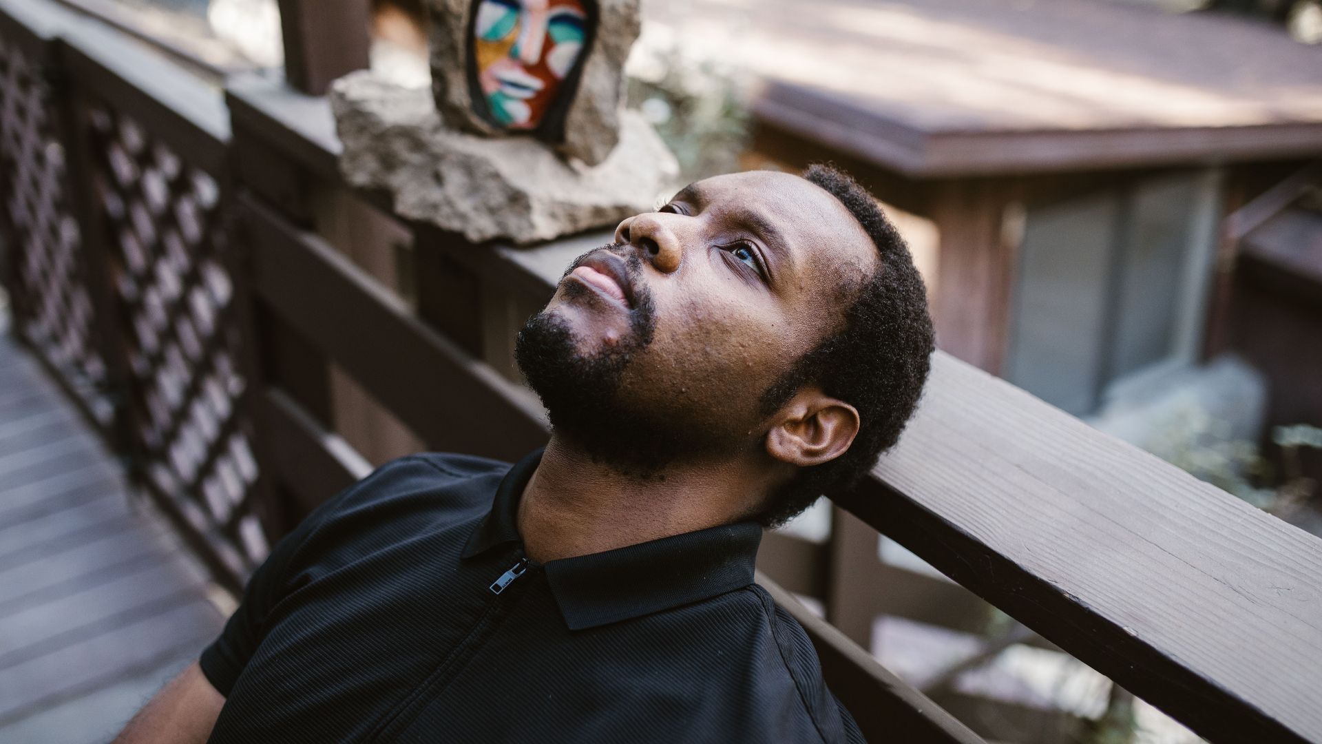 A man in thoughtful reflection sits outdoors against a wooden railing with abstract art behind.