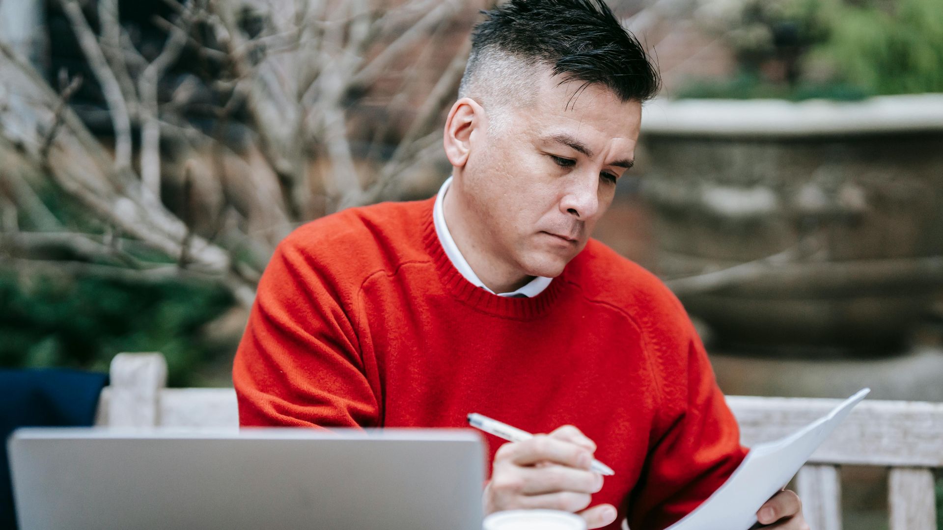 A man in a red sweater concentrating on paperwork while working remotely with a laptop outdoors.