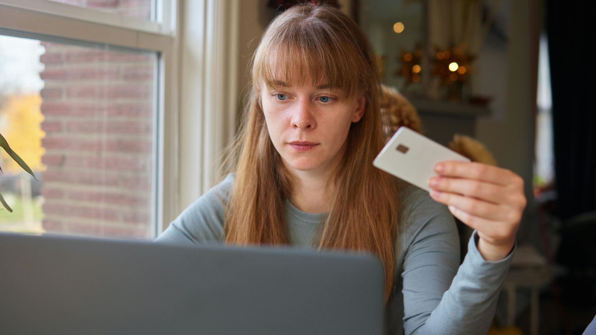 A young woman holds a credit card while using her laptop indoors.