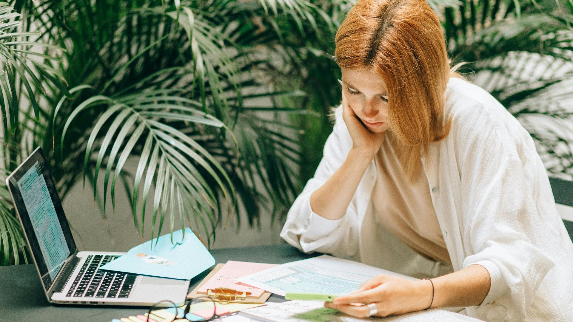 Woman in a thoughtful pose reviewing documents on a table with a laptop, surrounded by greenery.