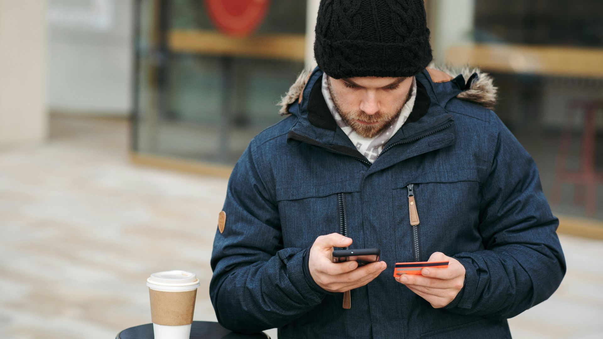 Concentrated young man in warm clothes and hat entering details of credit card while making online purchase via smartphone standing on city street with takeaway coffee