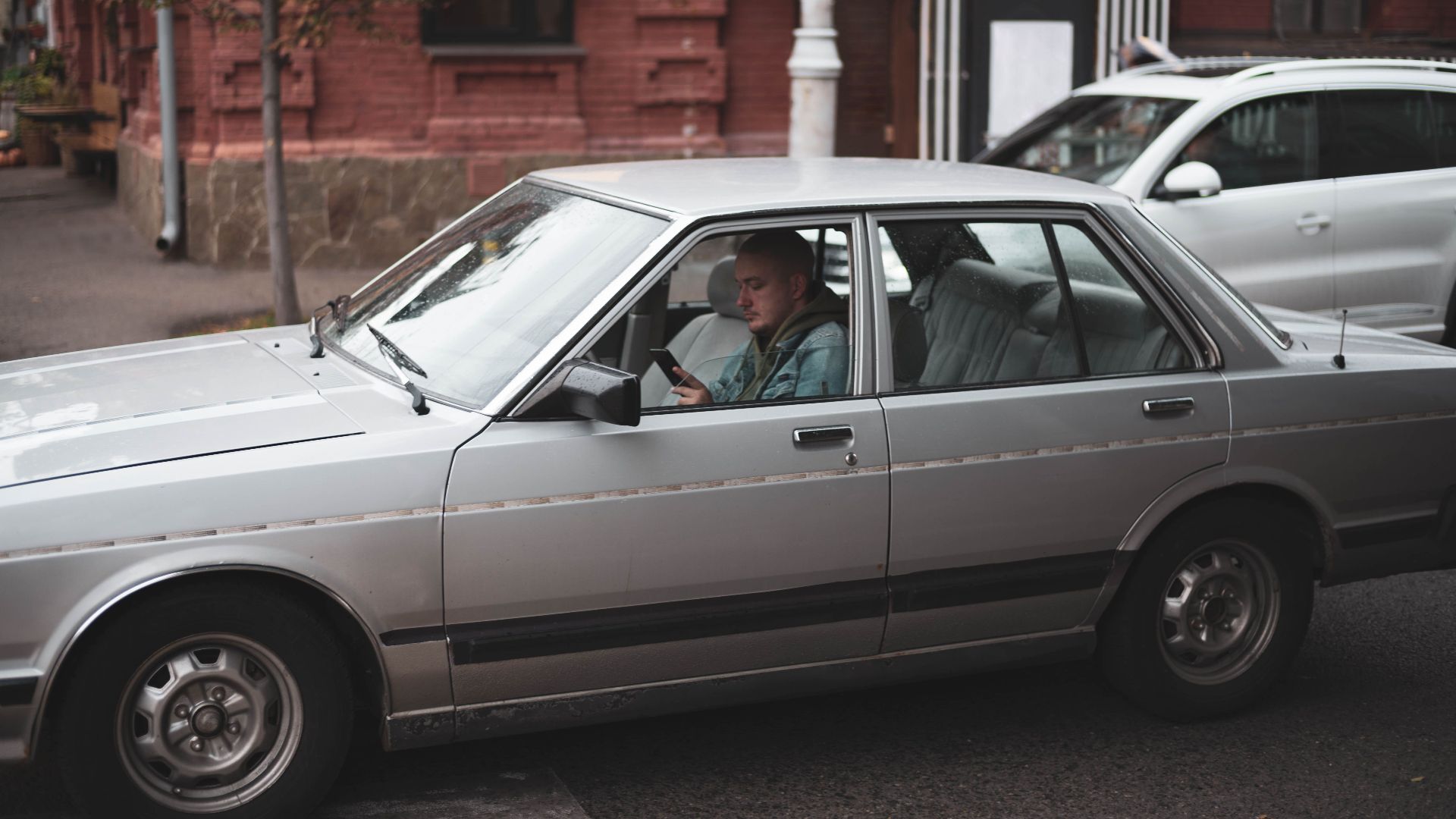 A classic silver sedan with a driver using a mobile phone in urban traffic.