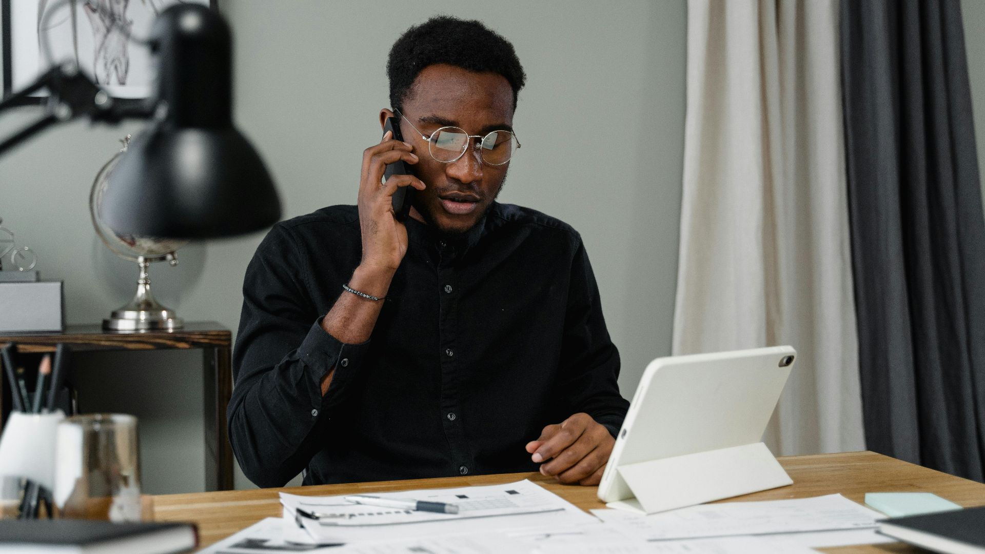 Focused man with eyeglasses making a work-related phone call at his desk with documents and a tablet.