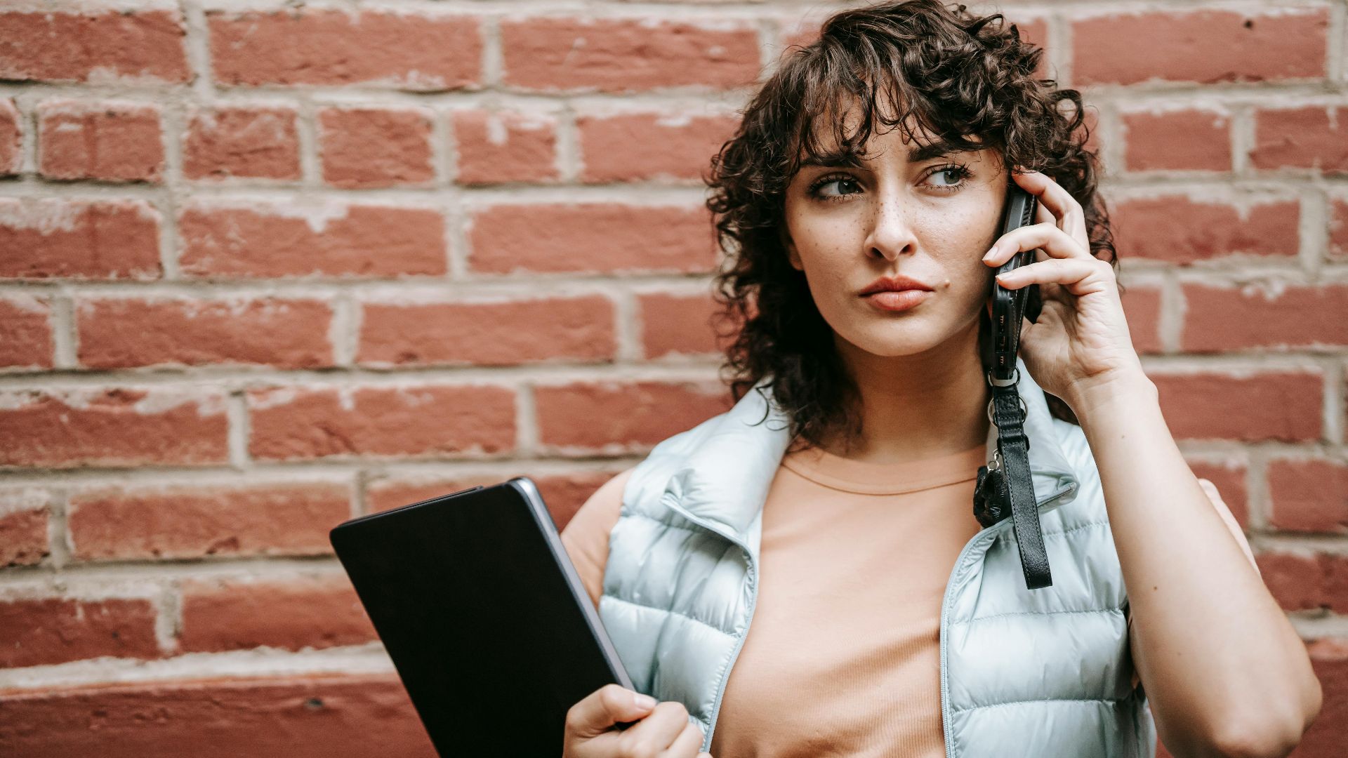 Adult attentive female remote worker with netbook talking on cellphone while looking away against brick wall