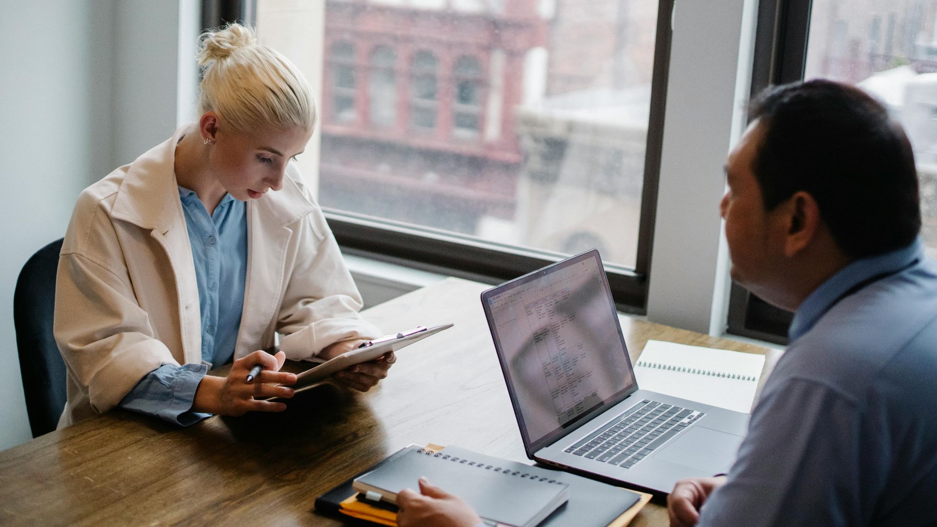From above of young female worker wearing modern coat analyzing details of project with colleague while sitting together at wooden desk with laptop and documents