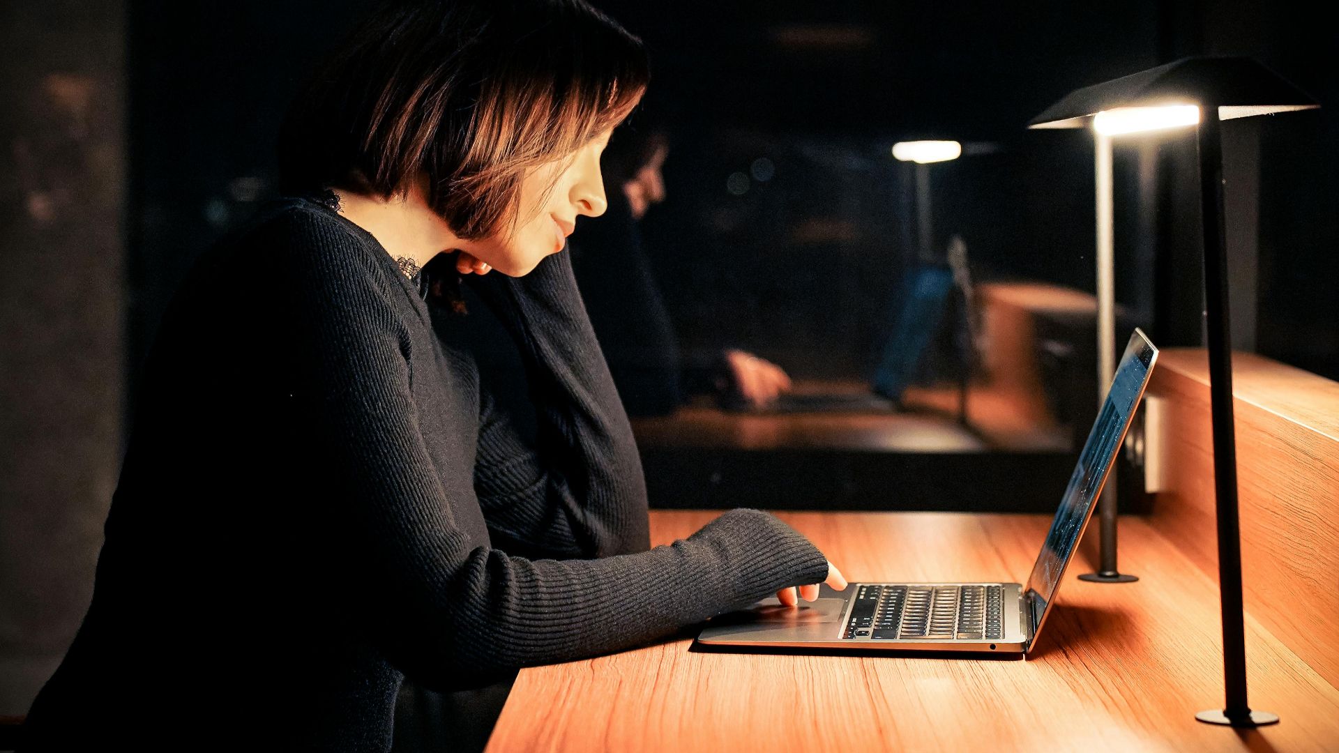 Focused woman using laptop at night in a dimly lit workspace, emphasizing concentration and technology.