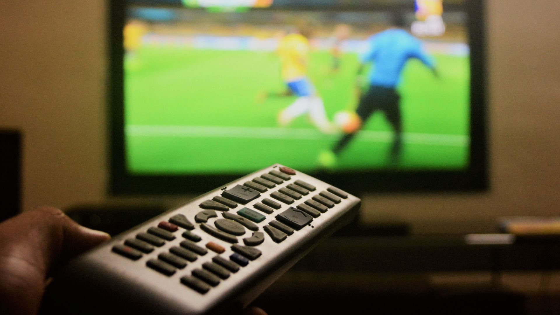 Close-up of a hand holding a remote control in front of a TV showing a football match.