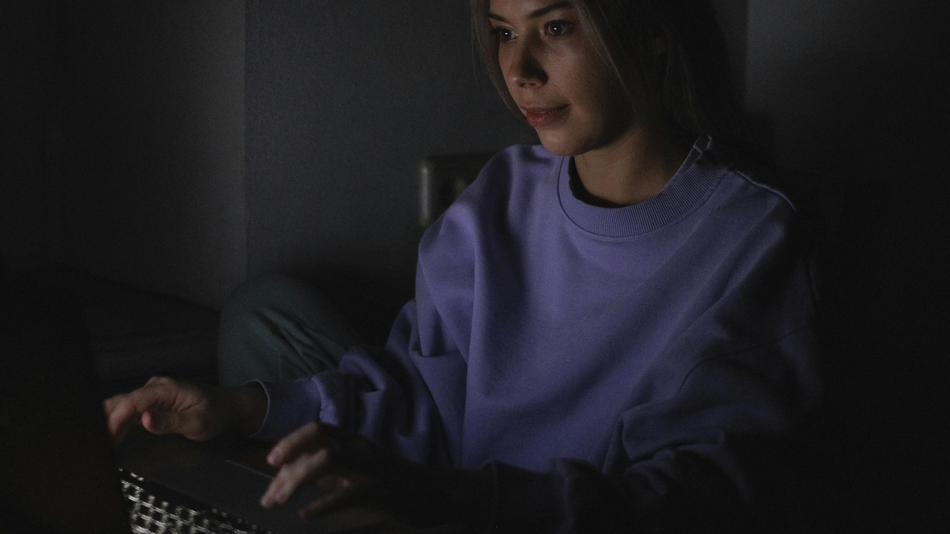 A woman wears a sweater and uses a laptop in a dark room, creating a focused and calm atmosphere.