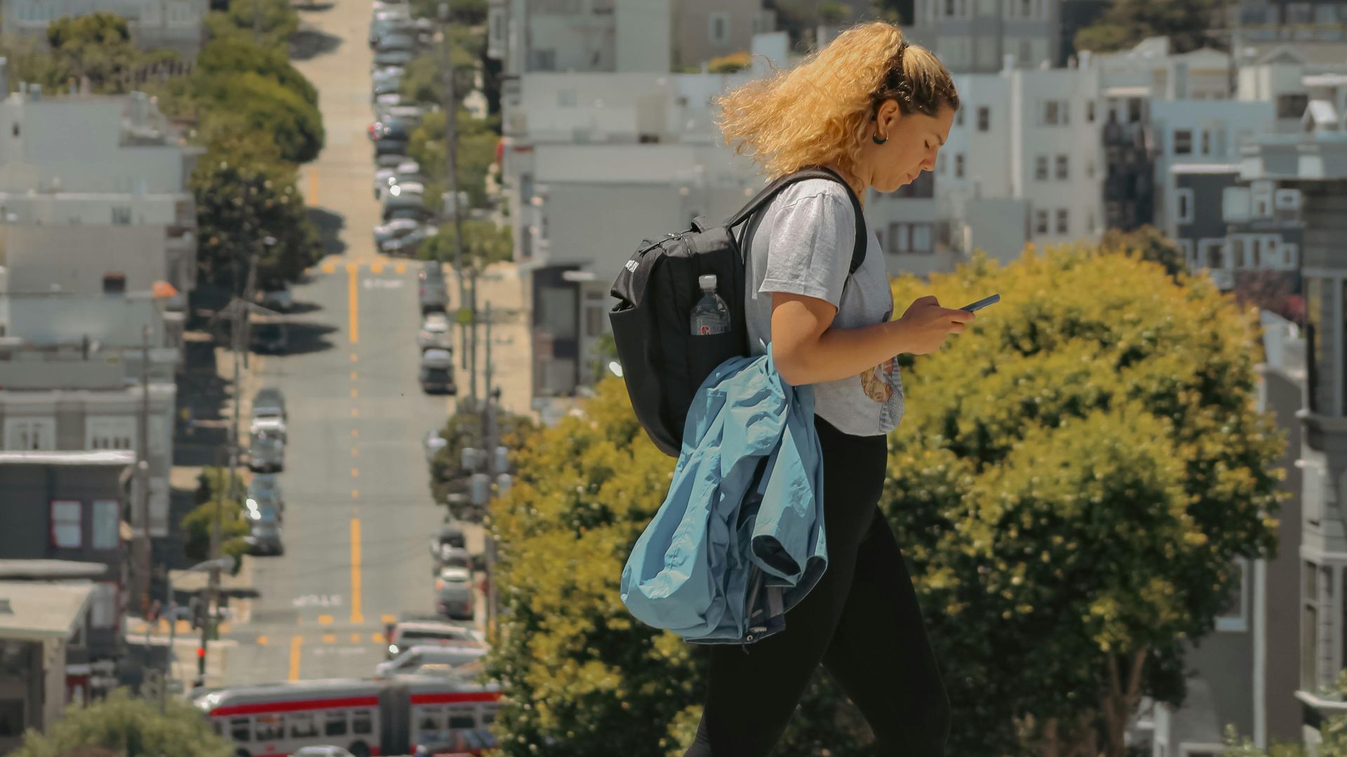 A woman with a backpack crossing a hilly street in San Francisco on a sunny day.