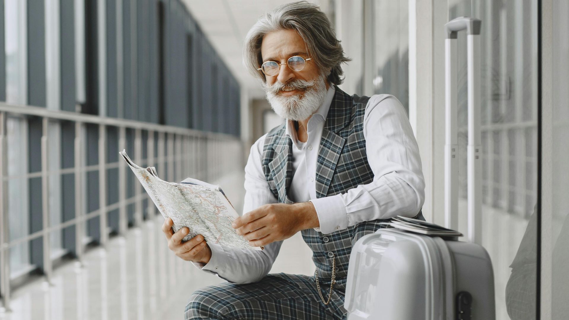 Senior man in airport hallway holding a map, sitting beside luggage, illustrating travel and exploration.