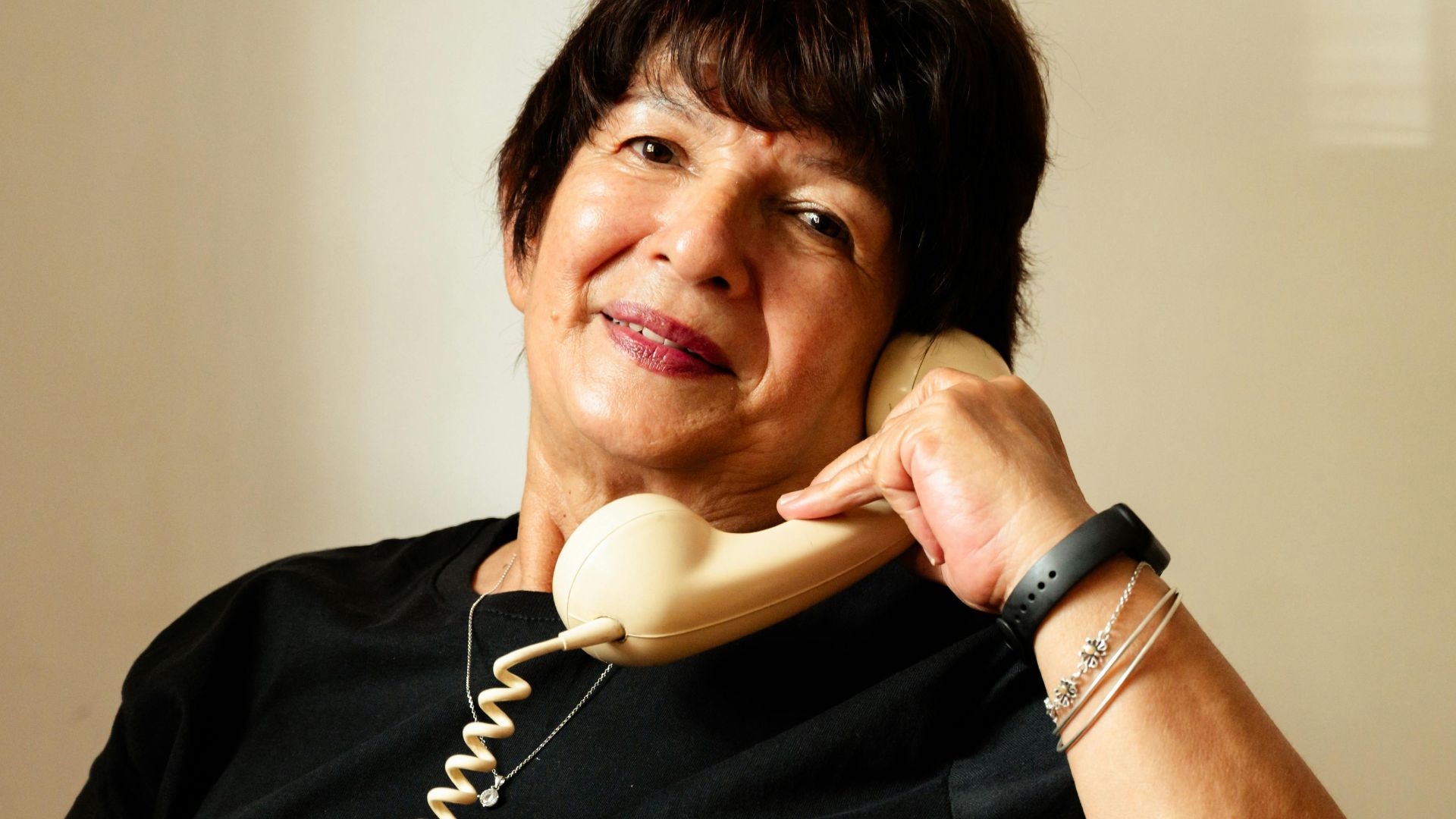 A senior woman wearing a black shirt smiles while talking on a vintage corded telephone indoors.