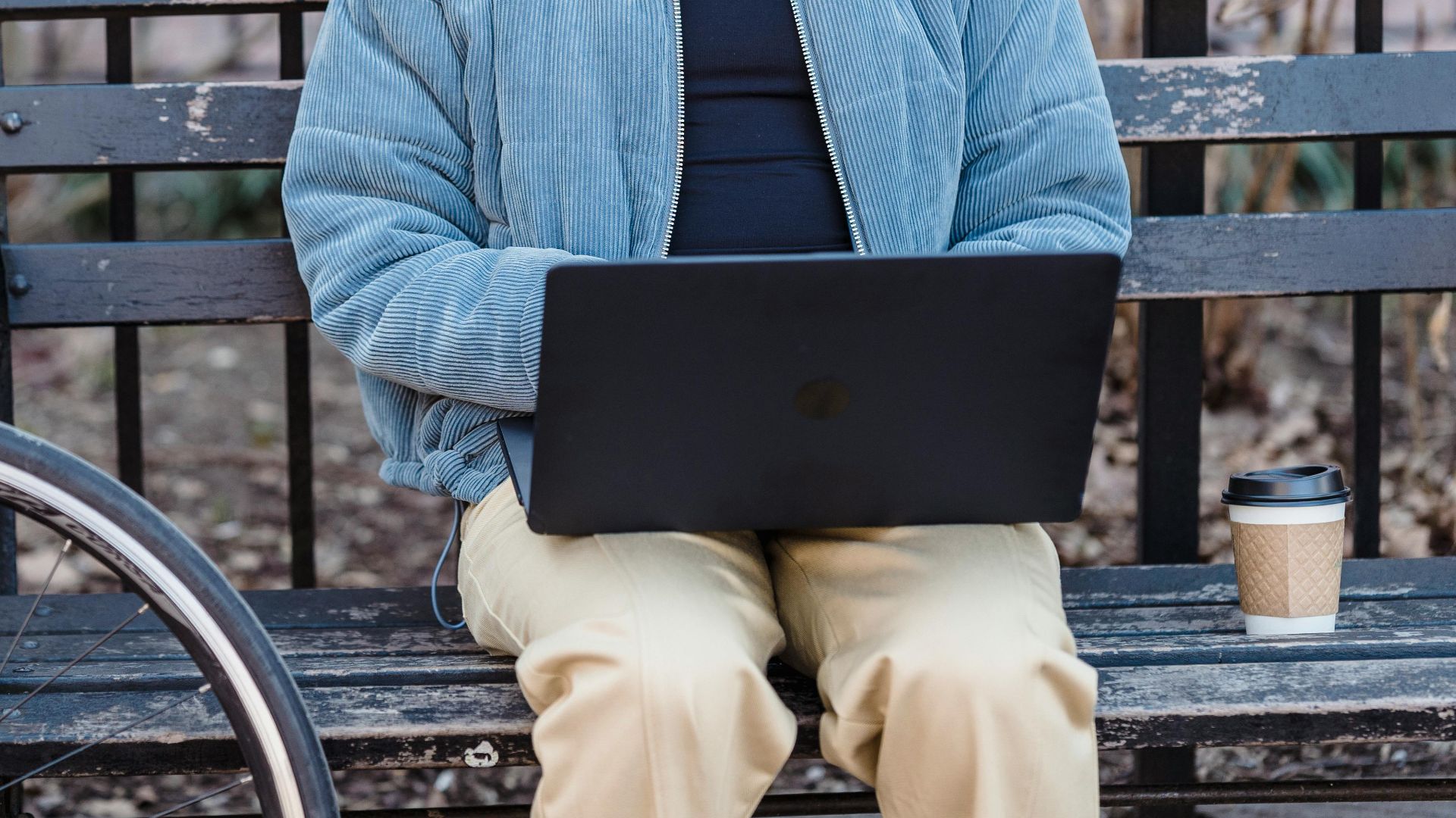 Focused young ethnic woman with curly brown hair in stylish warm jacket sitting on wooden bench in city park and typing on laptop during remote work