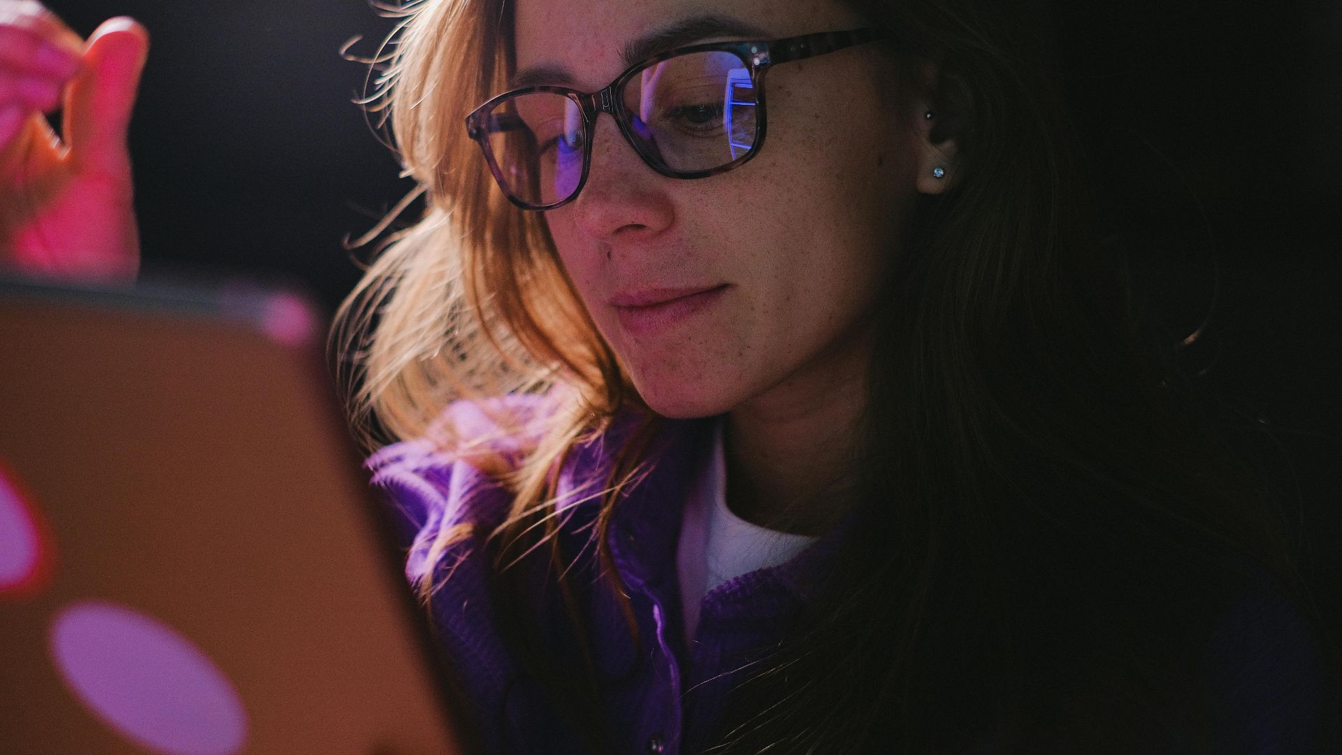 Concentrated female in eyeglasses wearing casual clothes browsing internet on modern tablet in room with low light during online work