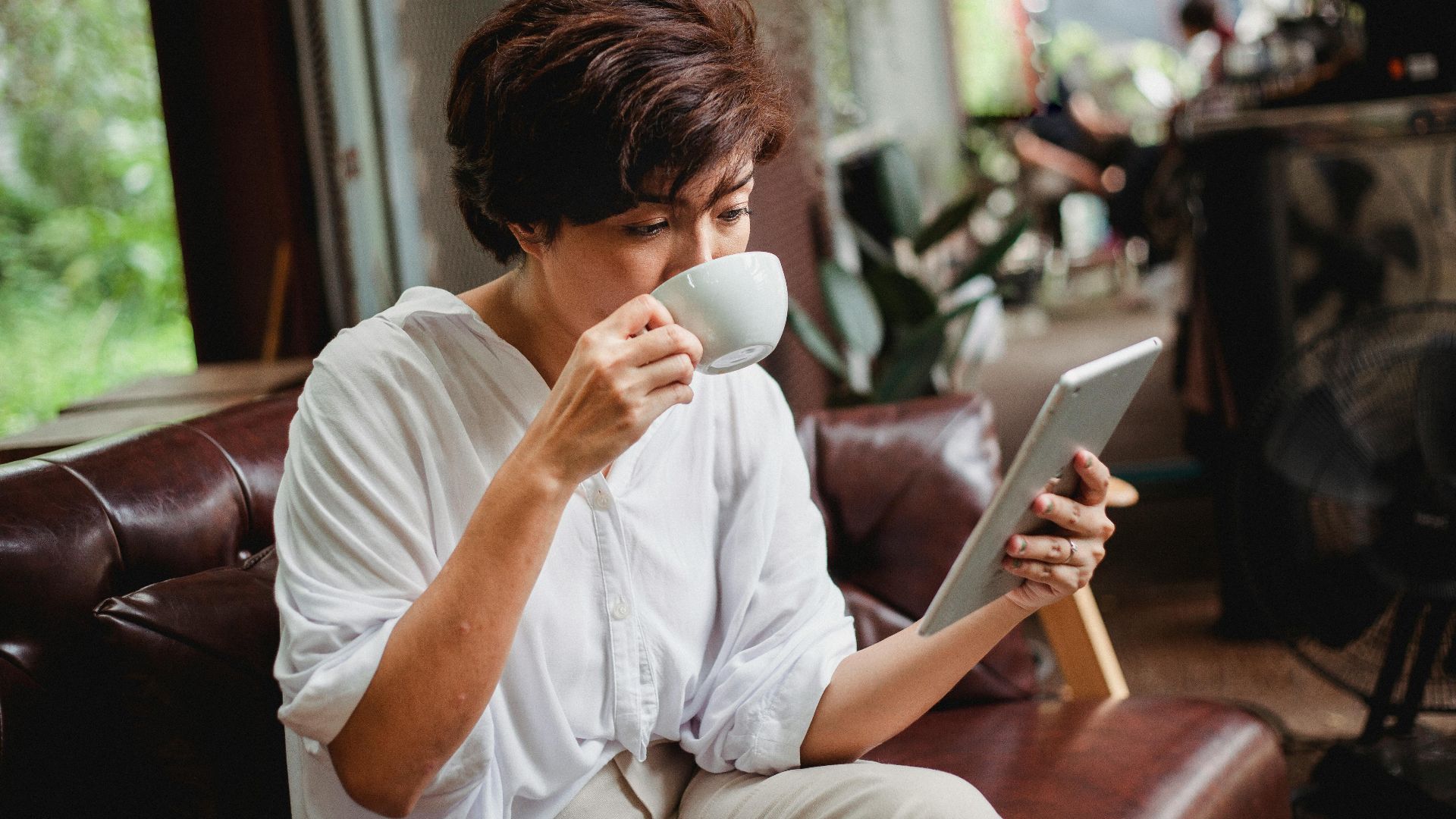 Focused Asian female in trendy clothes drinking cup of beverage and looking at screen of tablet