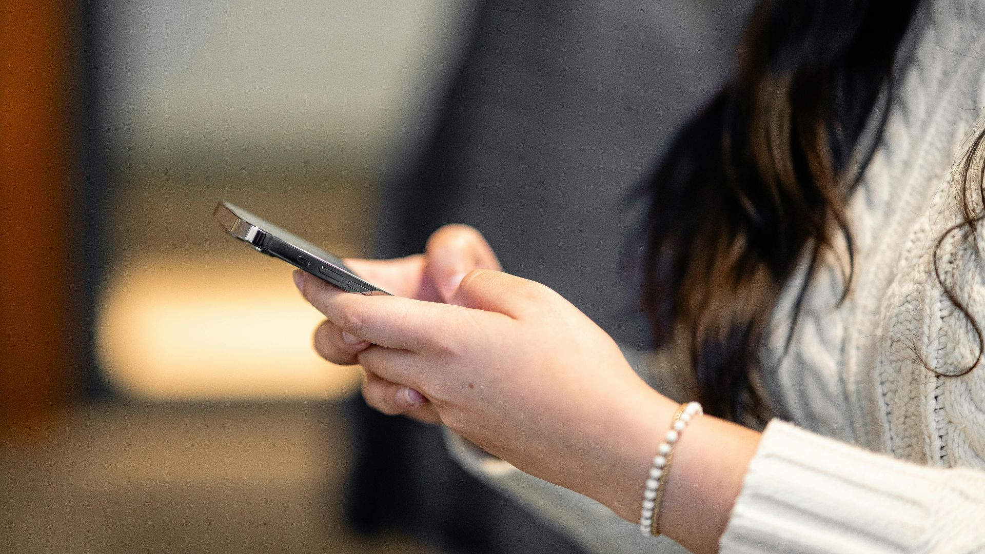 Close-up of a woman in a sweater holding a smartphone indoors.