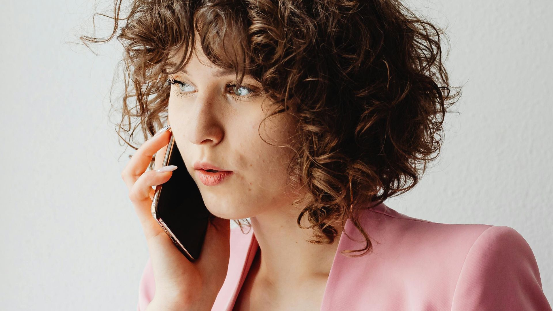 Confident businesswoman with curly hair talking on the phone indoors wearing a pink suit.