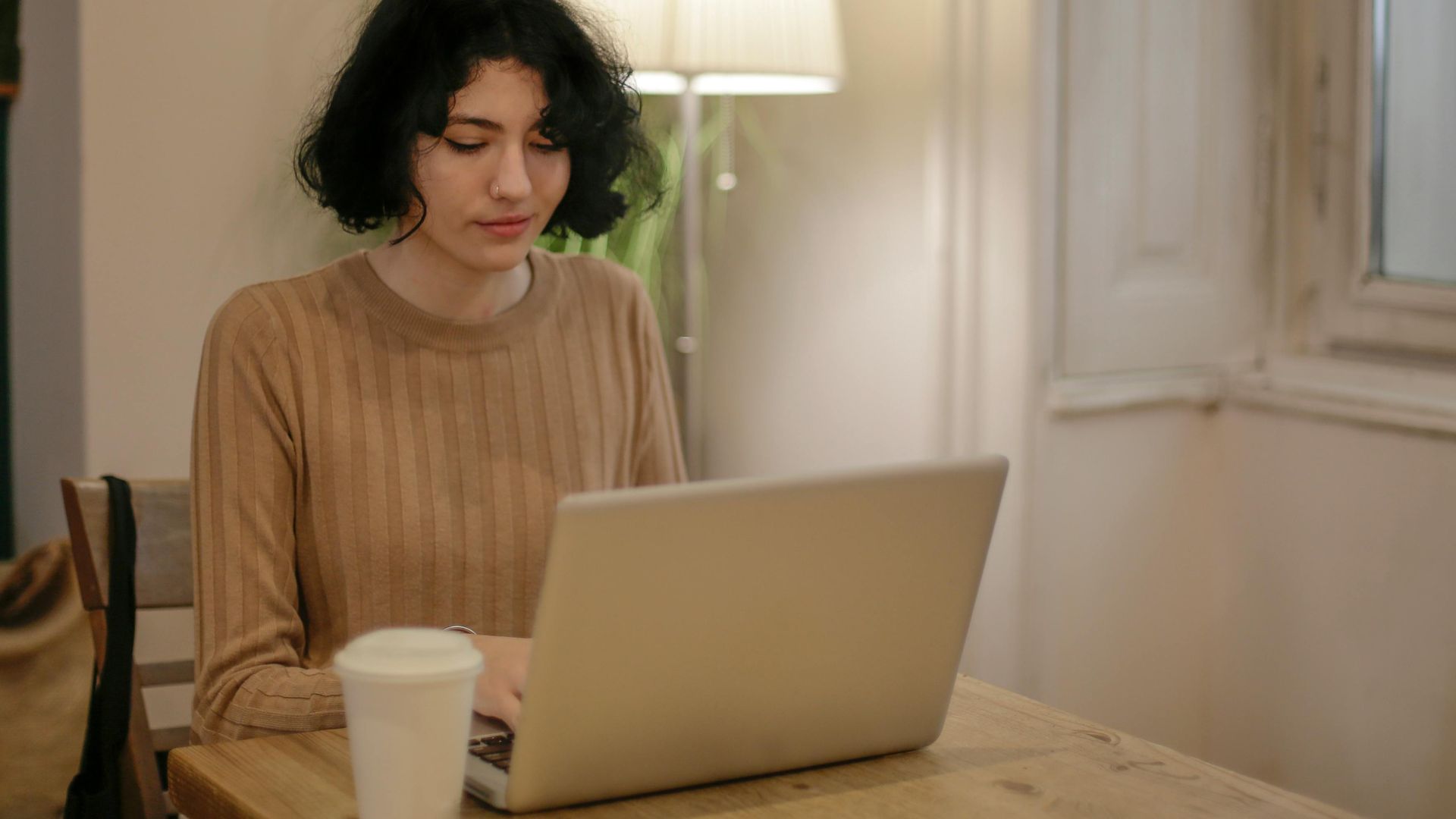 Woman sitting at a table using a laptop with a coffee cup, in a cozy indoor setting.