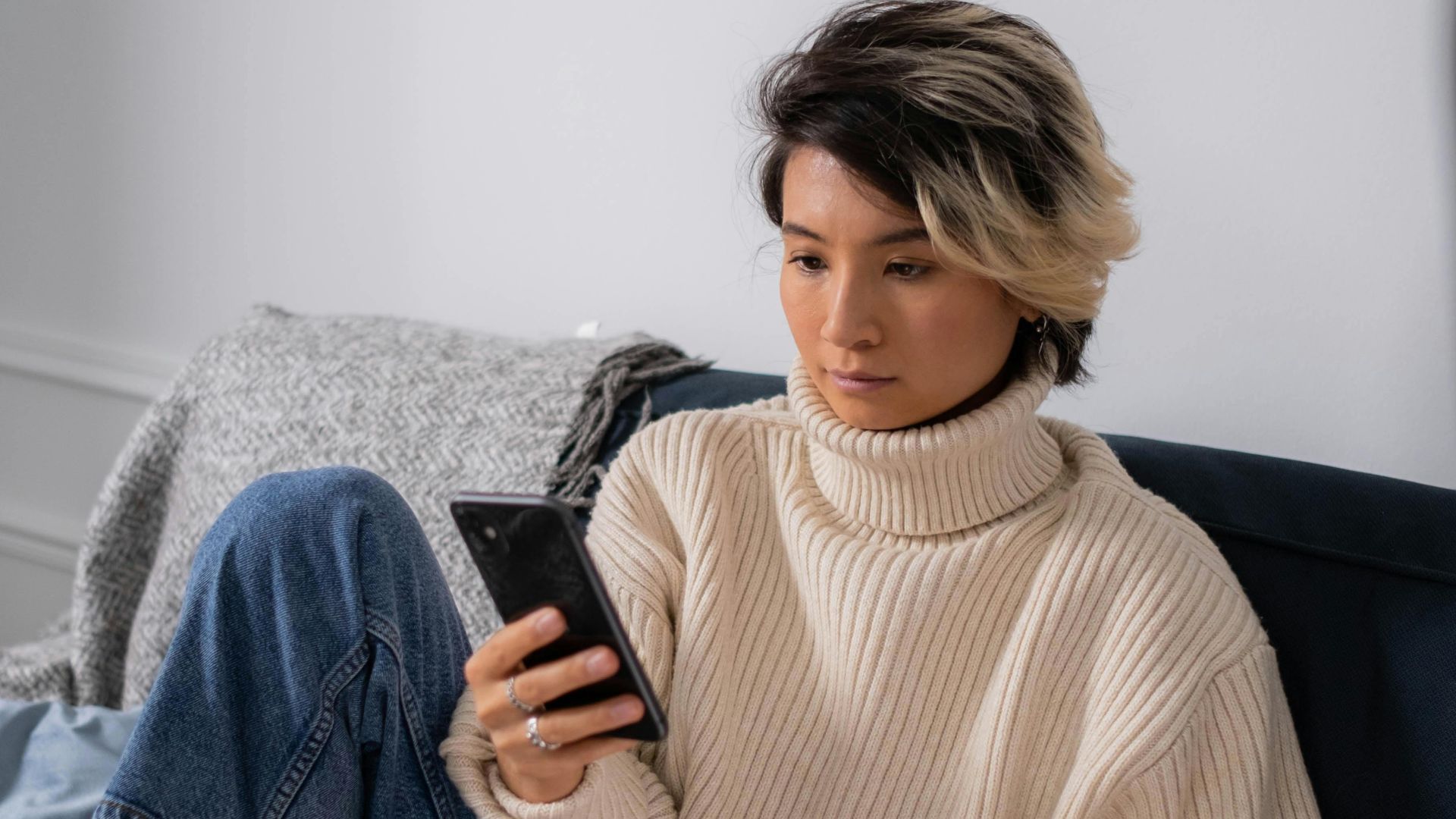 Asian woman in cozy sweater using smartphone and laptop on sofa indoors.