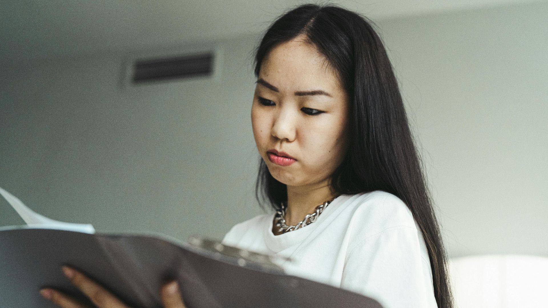 Asian woman in casual attire examining paperwork with concentration indoors.