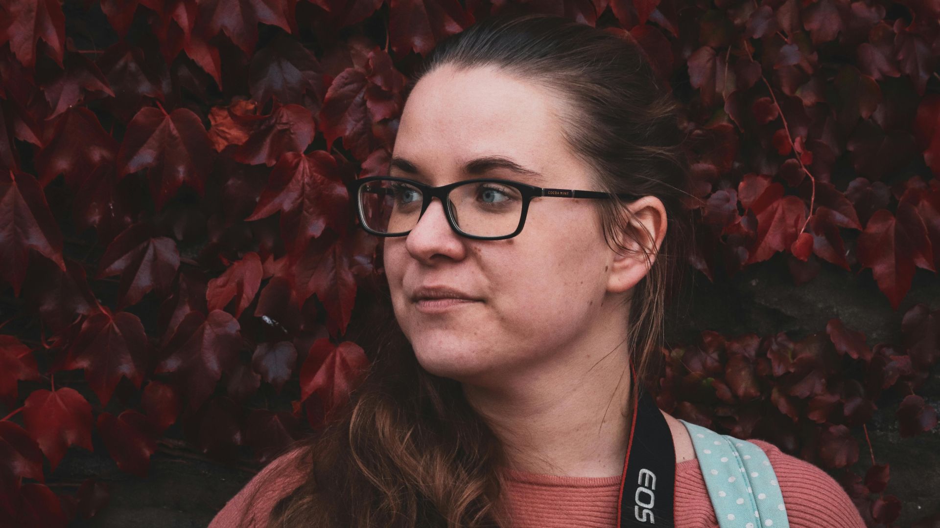 Portrait of a woman with eyeglasses standing against a vibrant red-leaf backdrop.