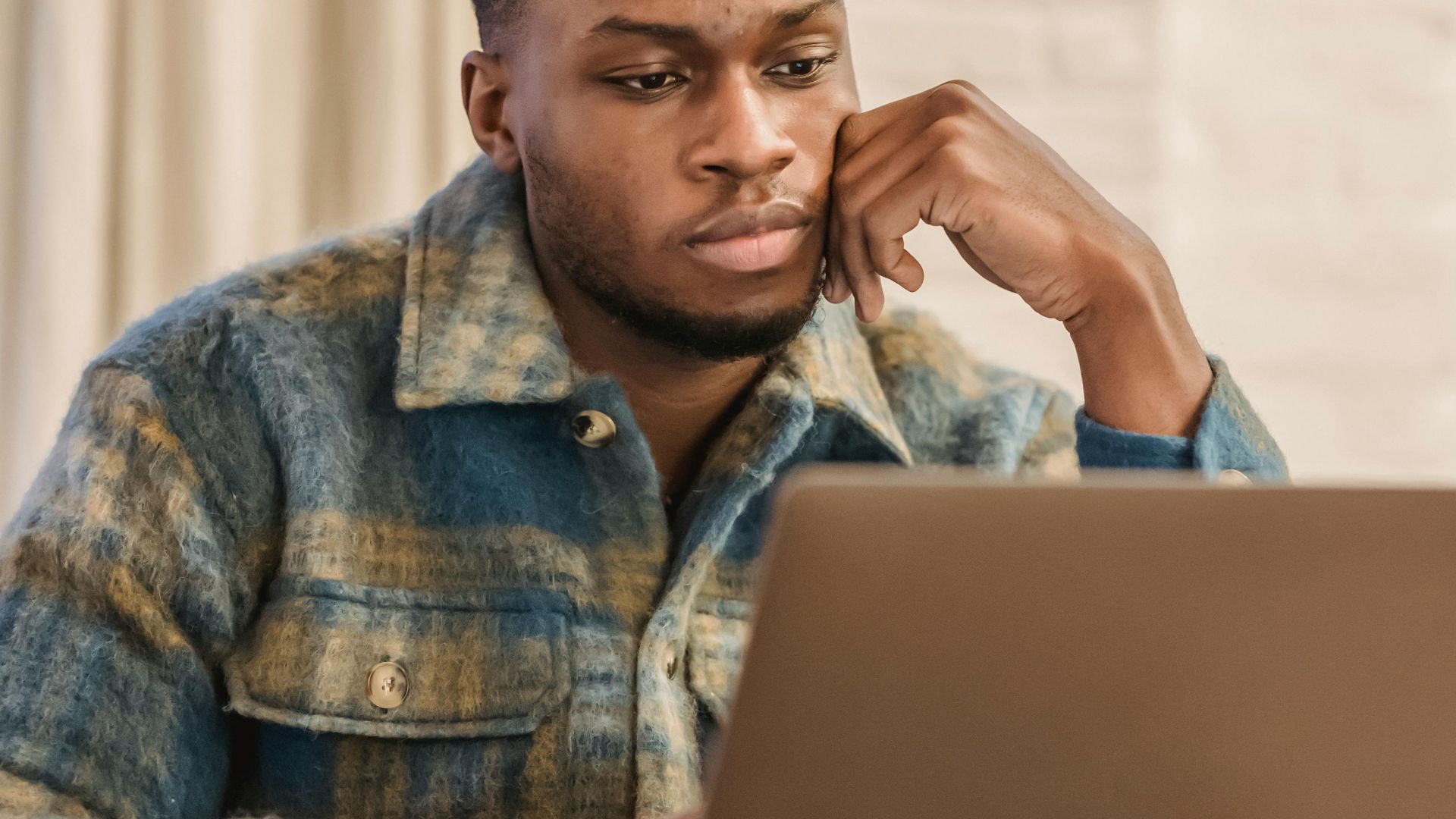 Thoughtful young African American male freelancer in casual clothes sitting at table with hand at chin and working distantly on laptop