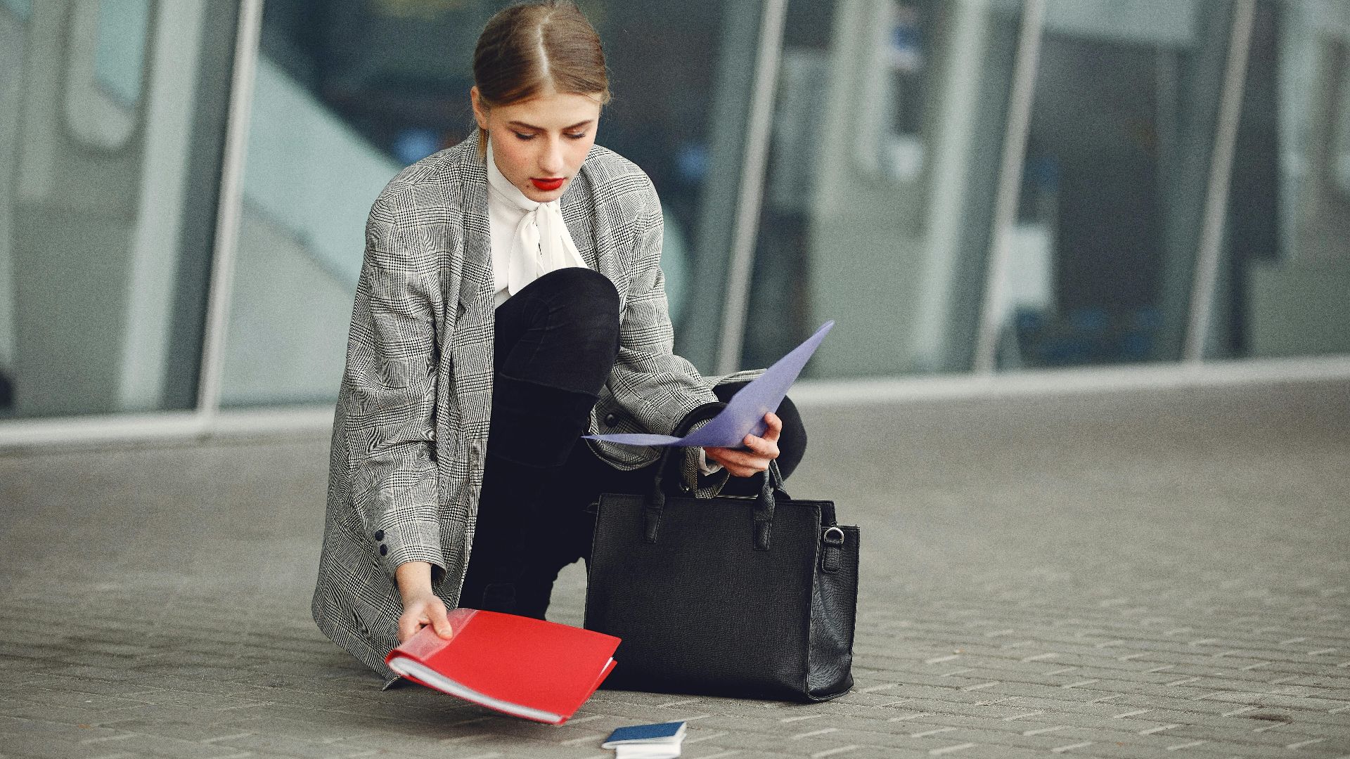 Distracted female manager in trendy wear collecting scattered documents and passport from sidewalk into briefcase near glass wall of modern building