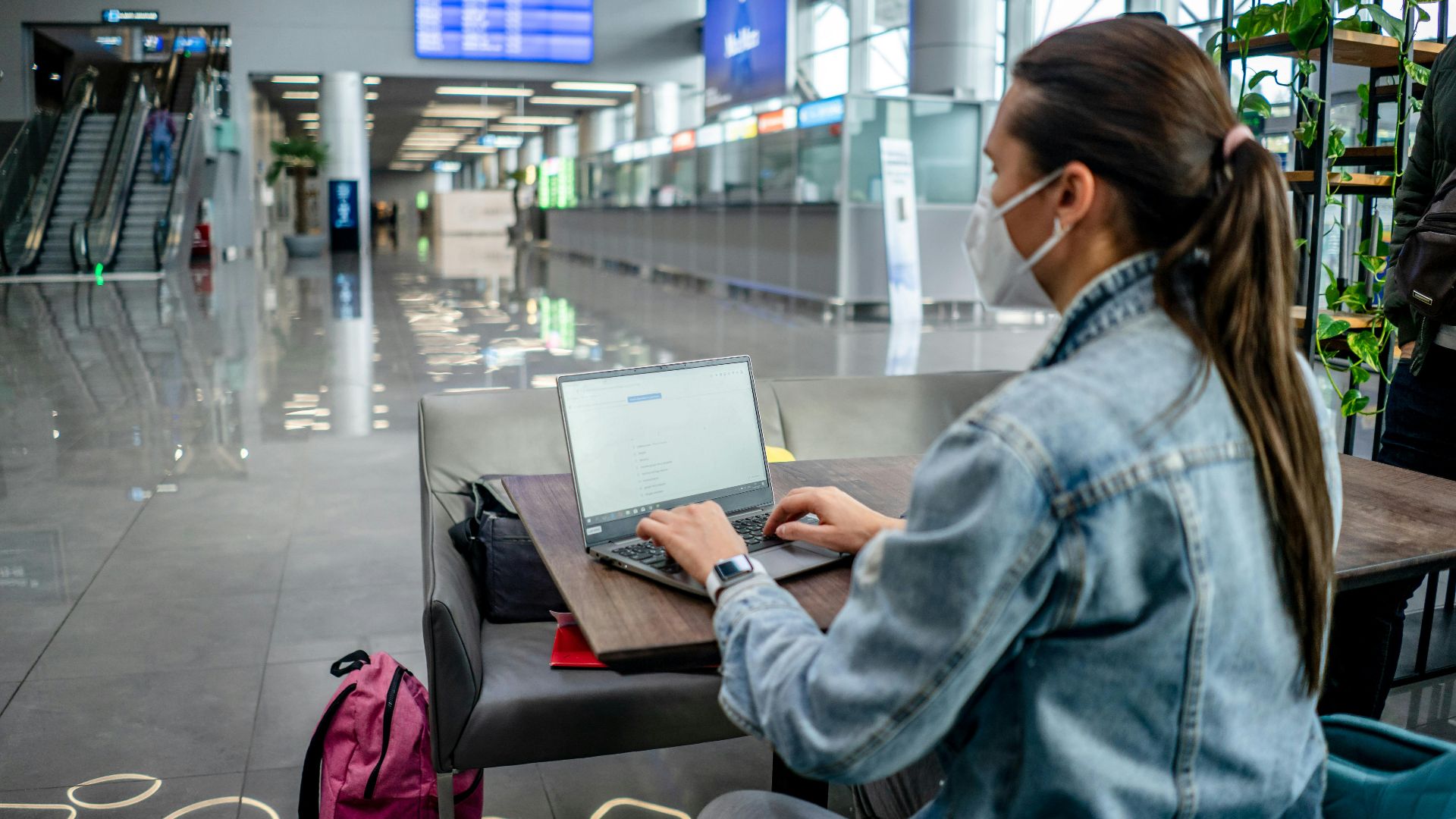 Side view of concentrated female freelancer in protective mask typing on netbook while sitting at table in modern airport terminal