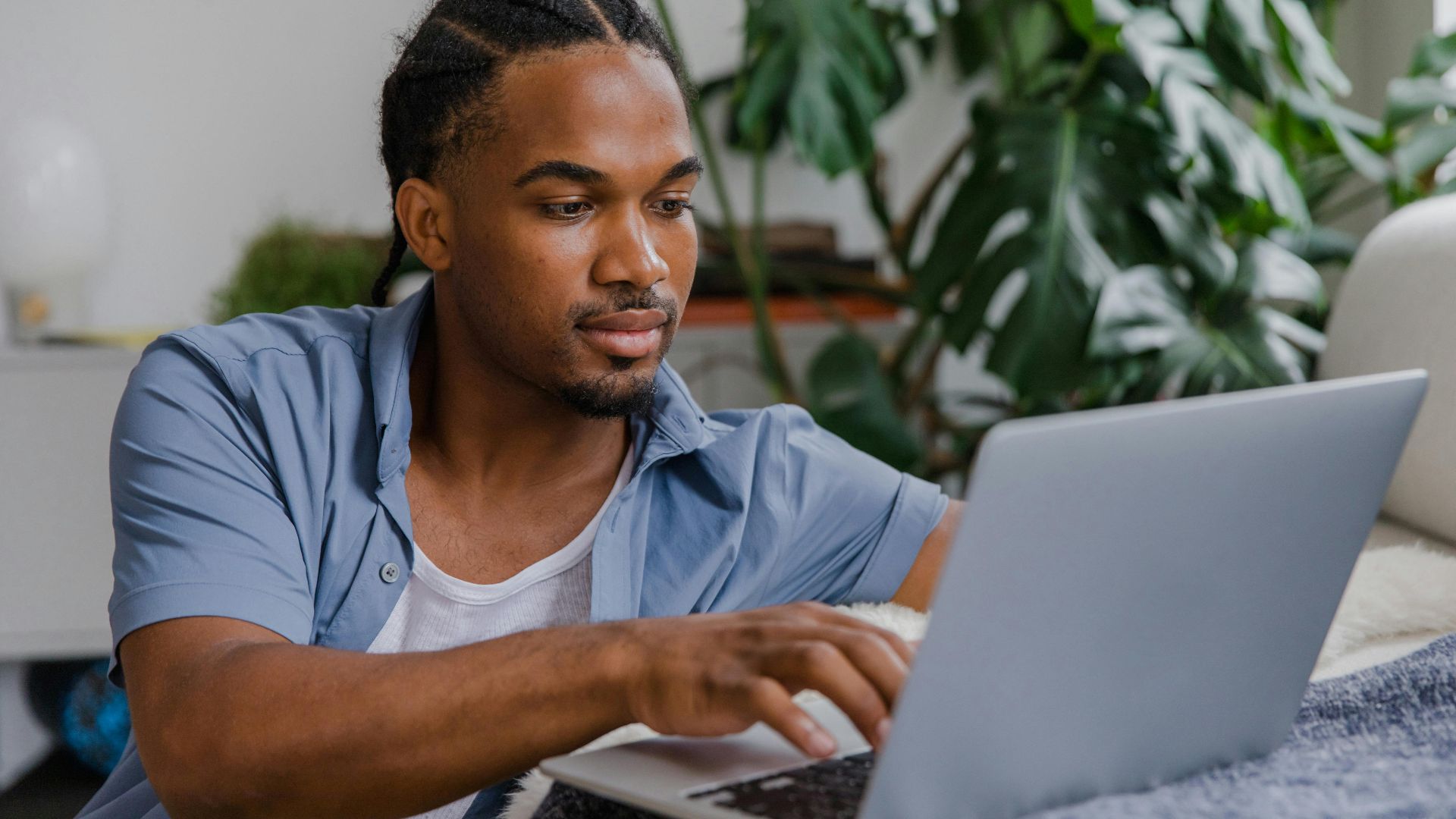 Young man in blue shirt working on laptop in a home office with a monstera plant.