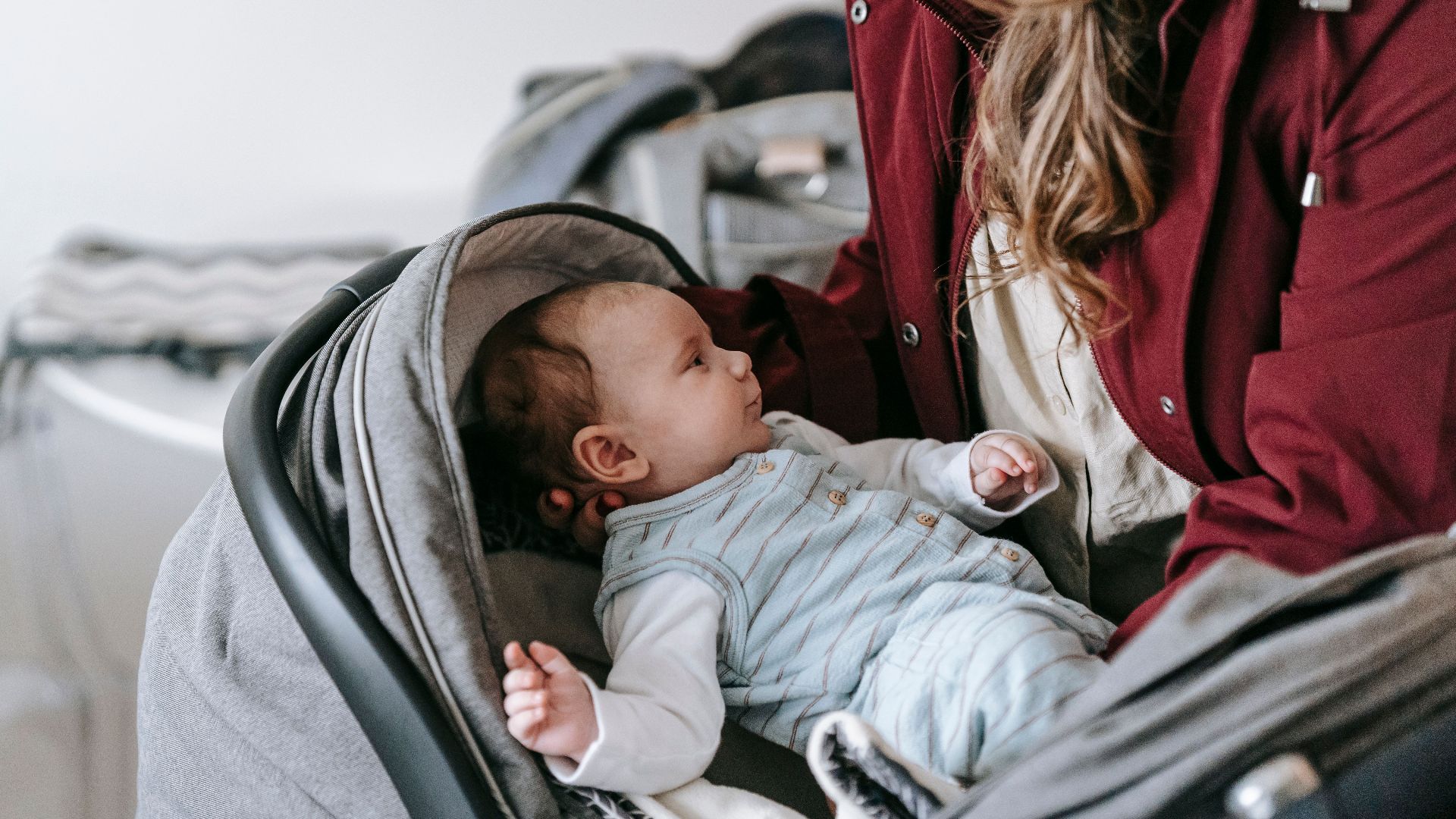 Crop anonymous caring mother in outerwear and hat putting infant baby into gray stroller while standing in light room at home