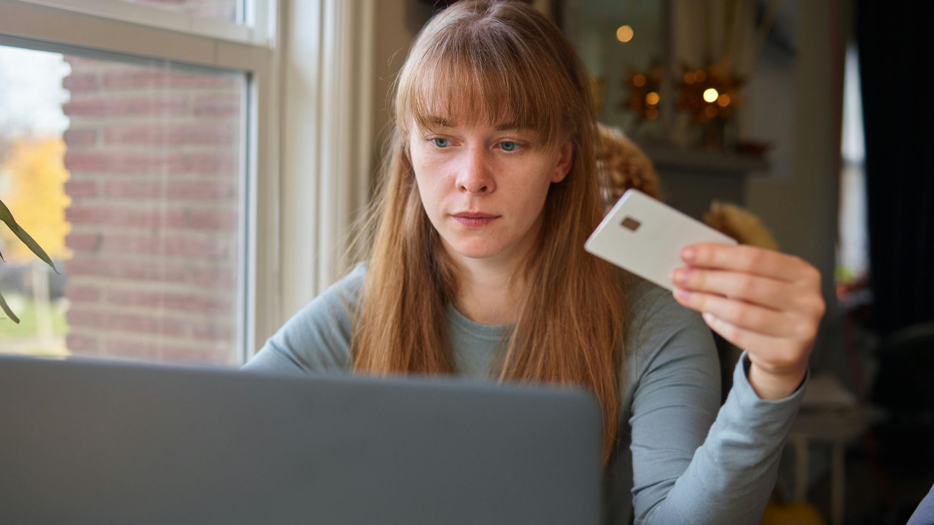A young woman holds a credit card while using her laptop indoors.