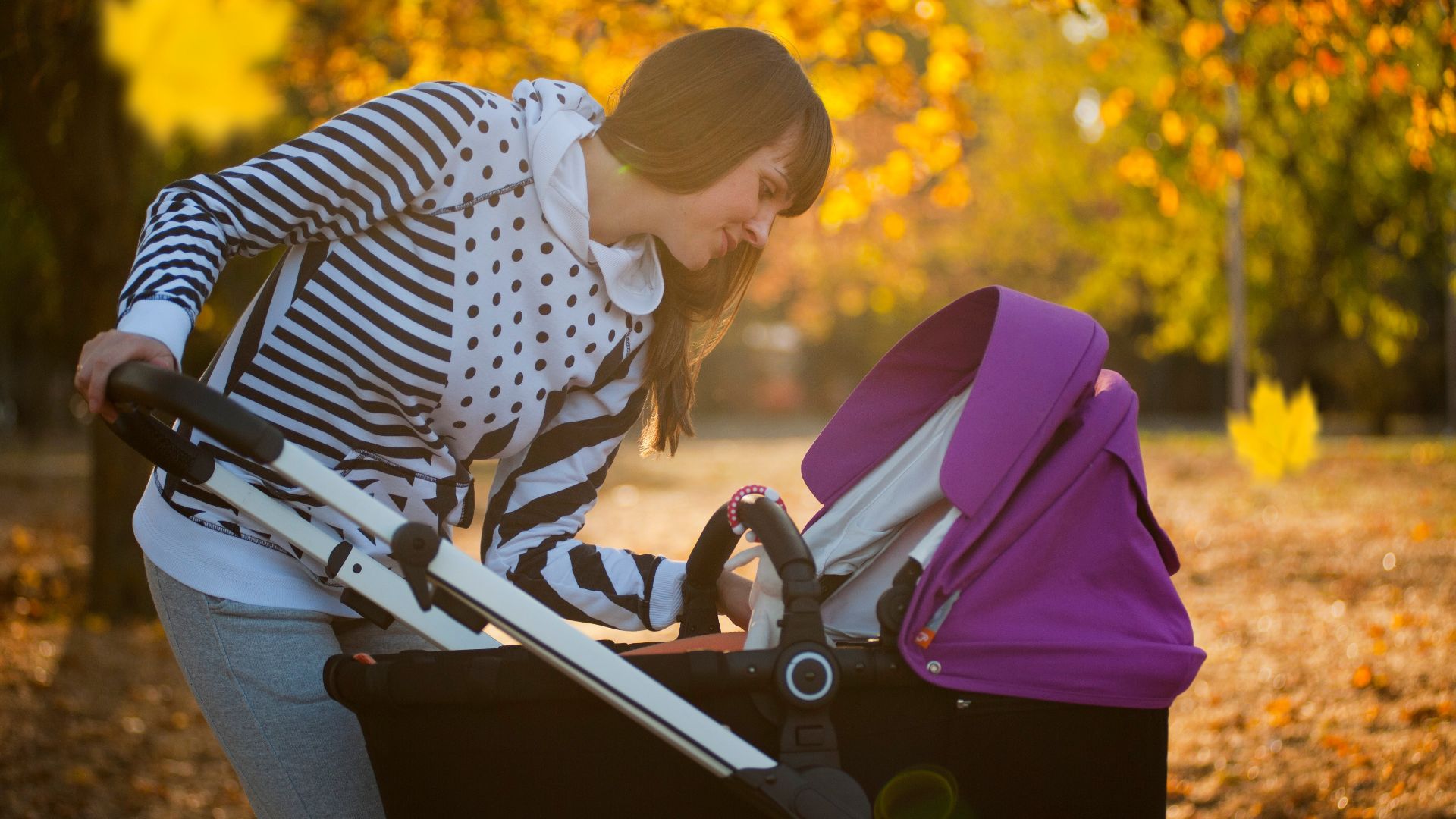 A mother smiling down at her baby in a stroller during an autumn stroll in a park.