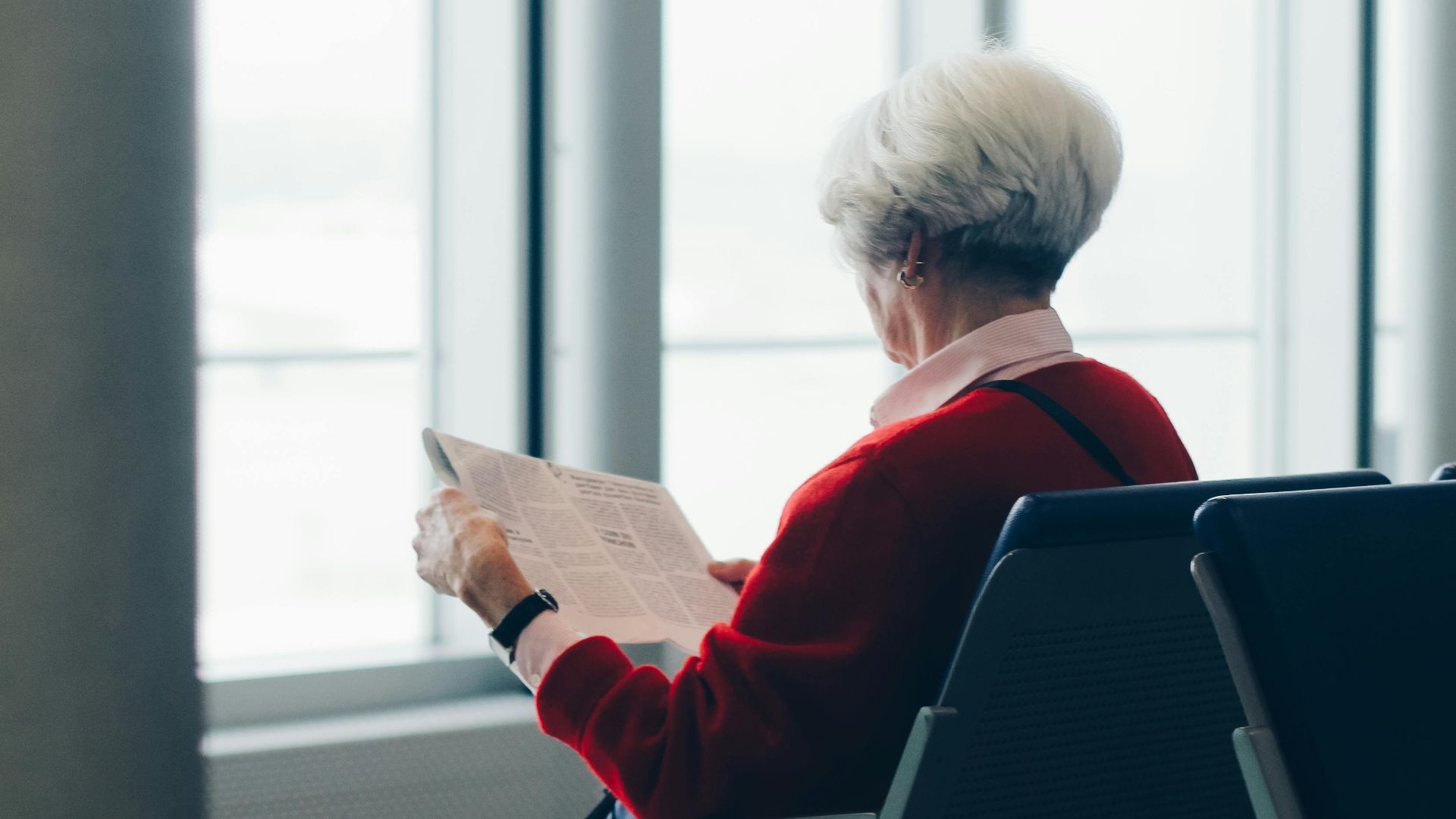 Elderly woman reading newspaper while seated at an airport gate.