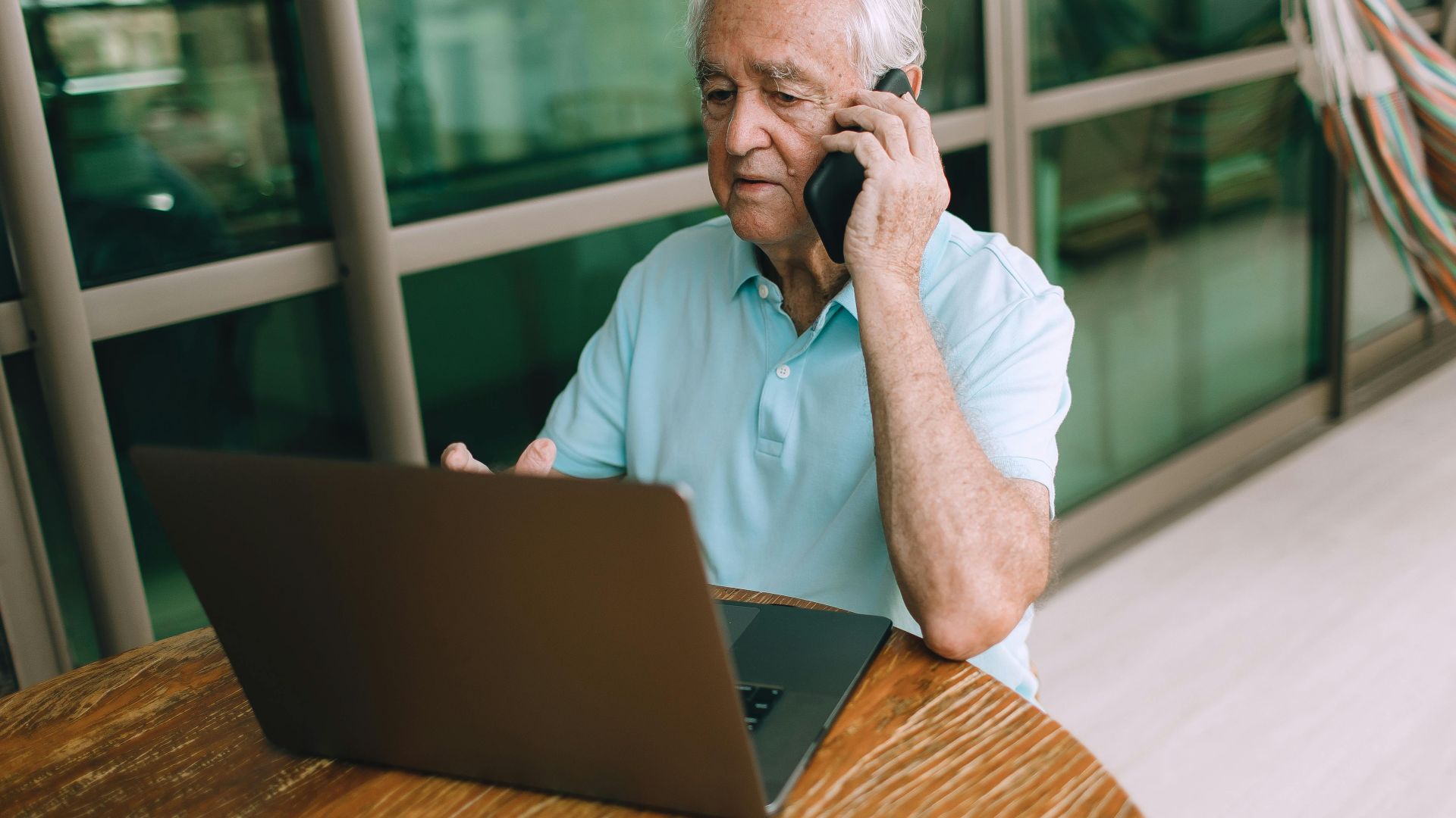 Elderly man multitasking with smartphone and laptop at home.